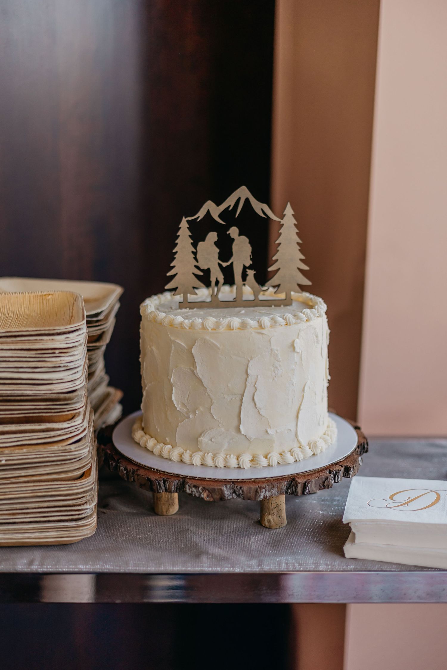 White wedding cake with woodland-themed topper displayed on rustic wood slice stand.