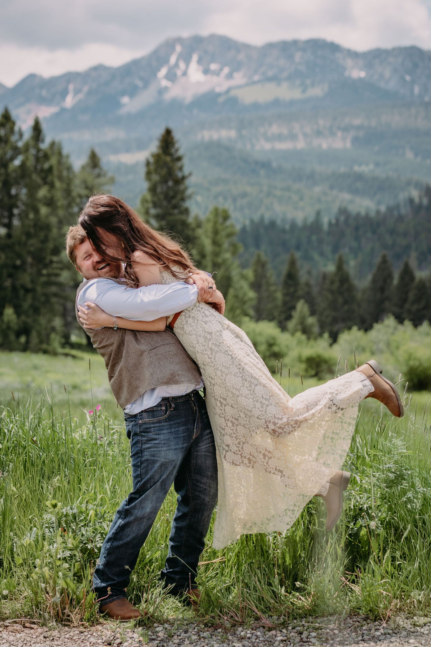A romantic embrace in a mountain meadow with evergreen forest and snow-capped peaks in the background.