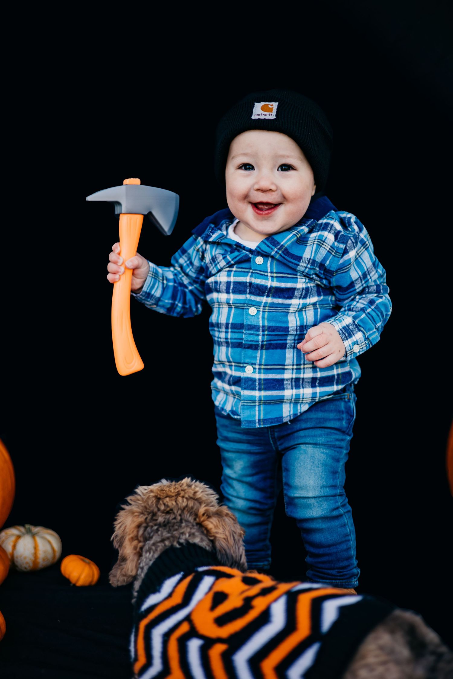Toddler in blue plaid shirt and beanie smiles while holding toy hammer near pumpkins.