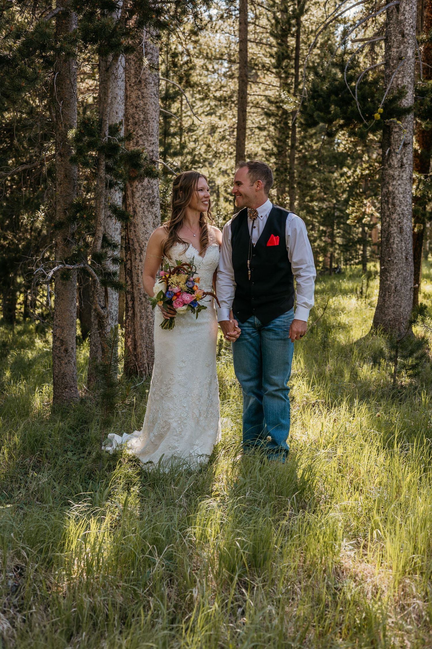 A couple walks through a sunlit forest in wedding attire surrounded by tall pine trees.