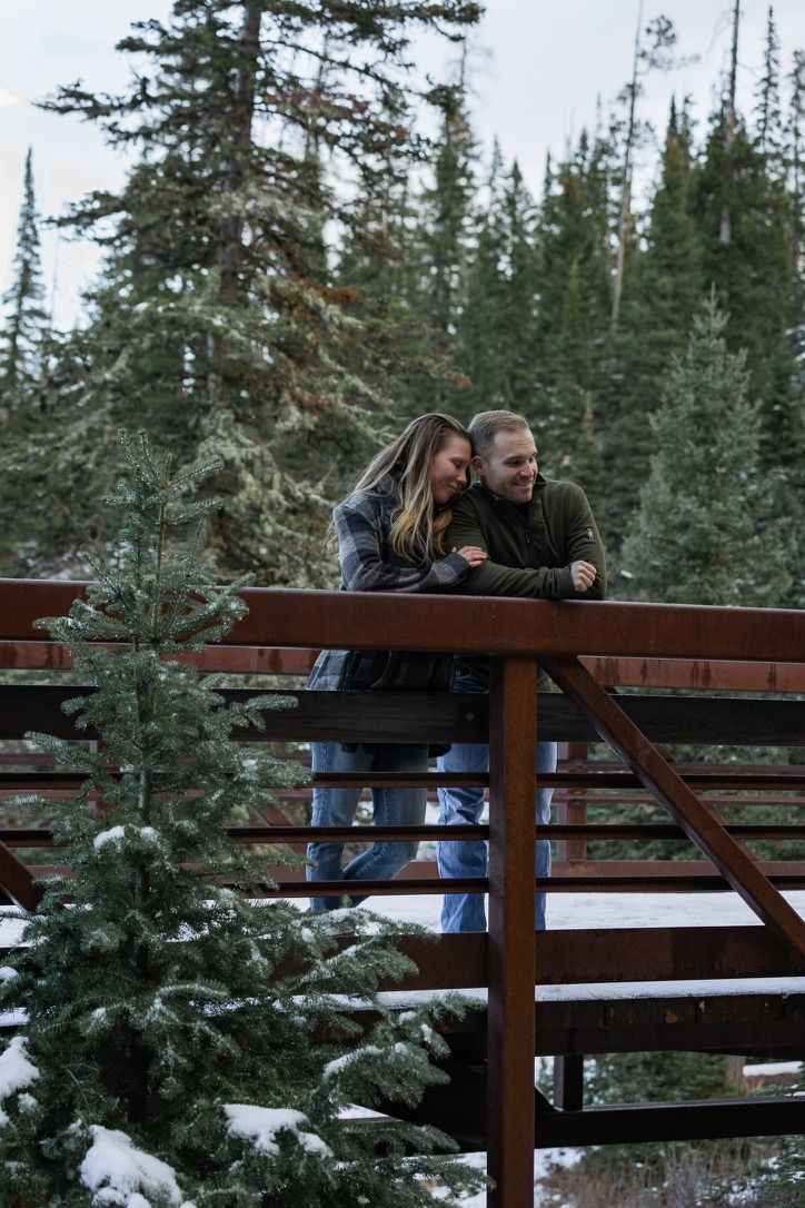 Two people share a tender moment on a wooden bridge surrounded by snow-dusted evergreen trees in winter.