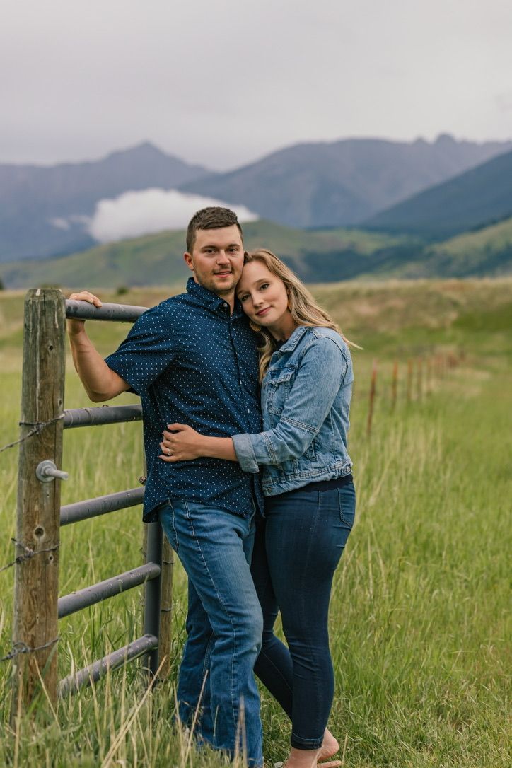 A couple leans against a wooden fence in a green field with mountain views in the background.