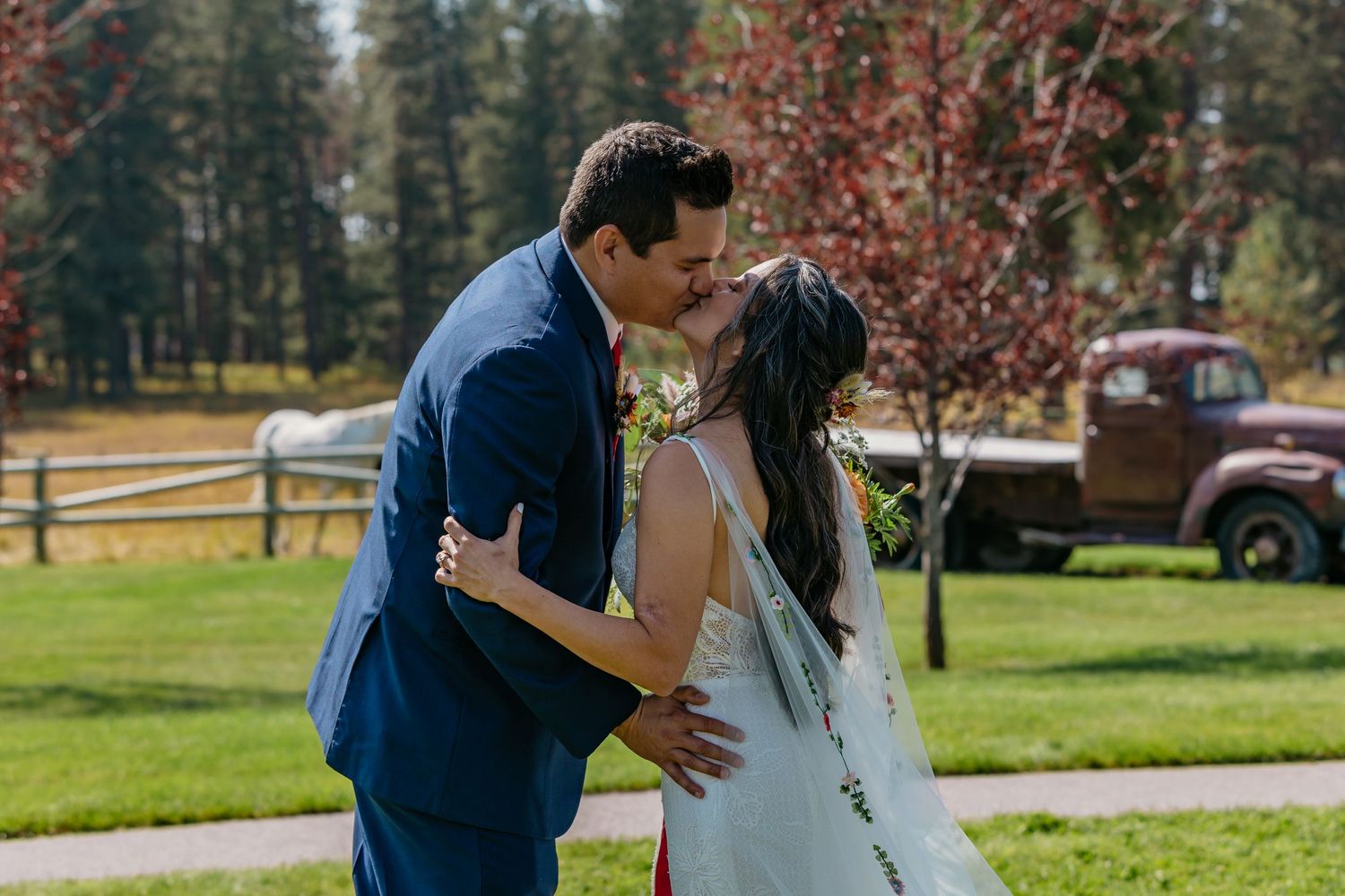 A couple shares a romantic kiss outdoors on their wedding day with a rustic ranch setting and vintage car in the background.