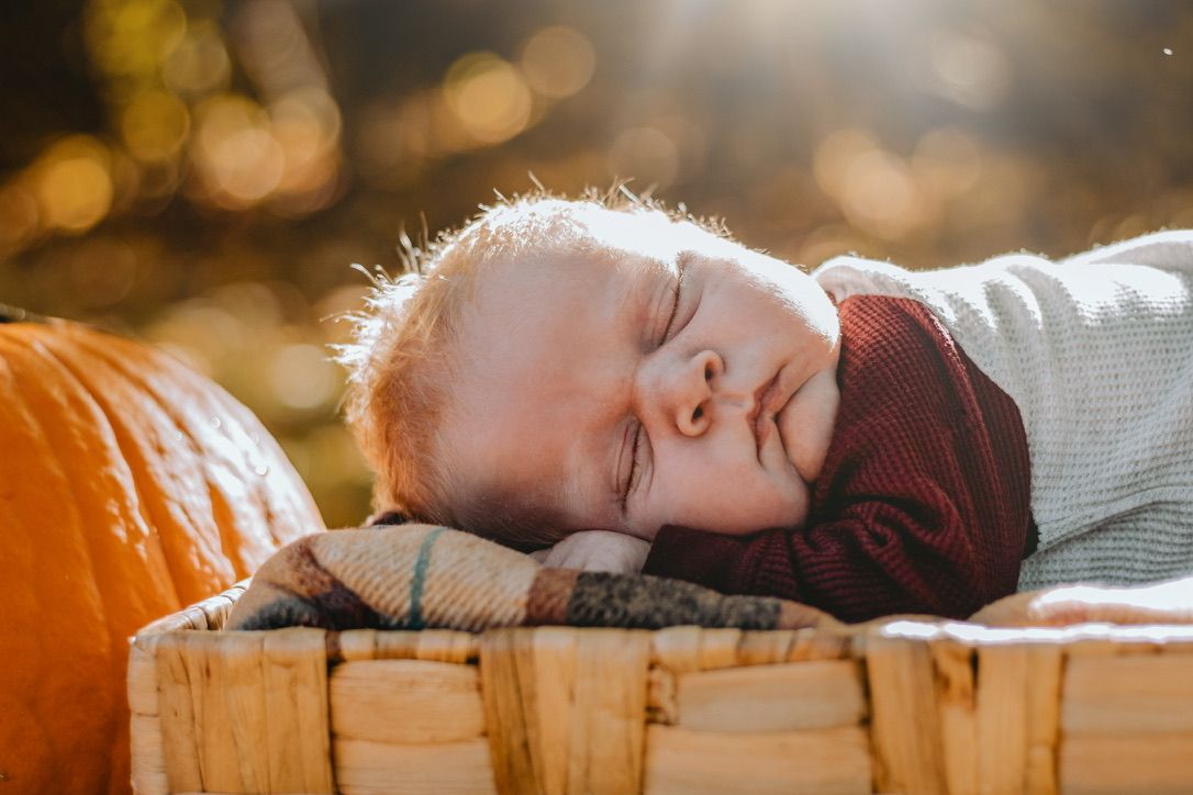 Sleeping baby rests head on plaid blanket next to orange pumpkin in autumn setting.