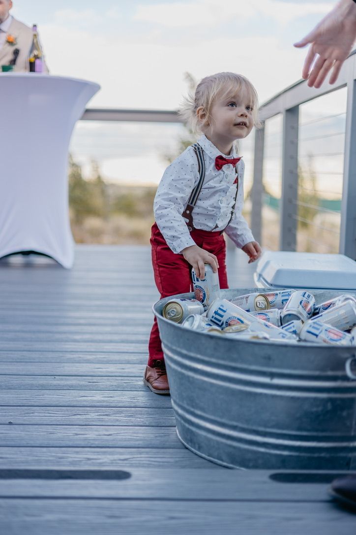 Small child in red overalls and bow tie reaches into metal tub filled with beverages at outdoor event.