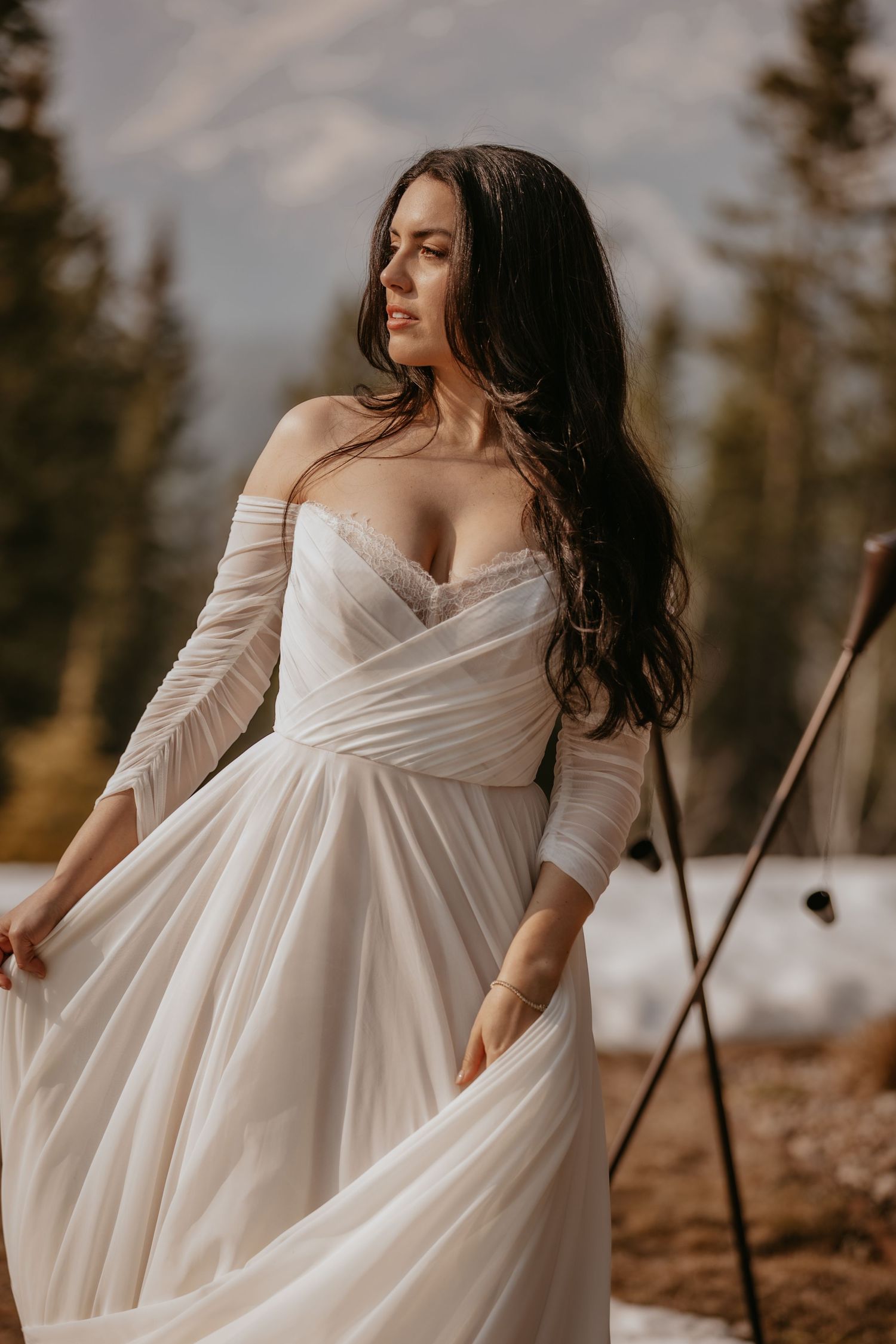 Bride in flowing off-shoulder wedding dress poses against mountain backdrop at sunset.