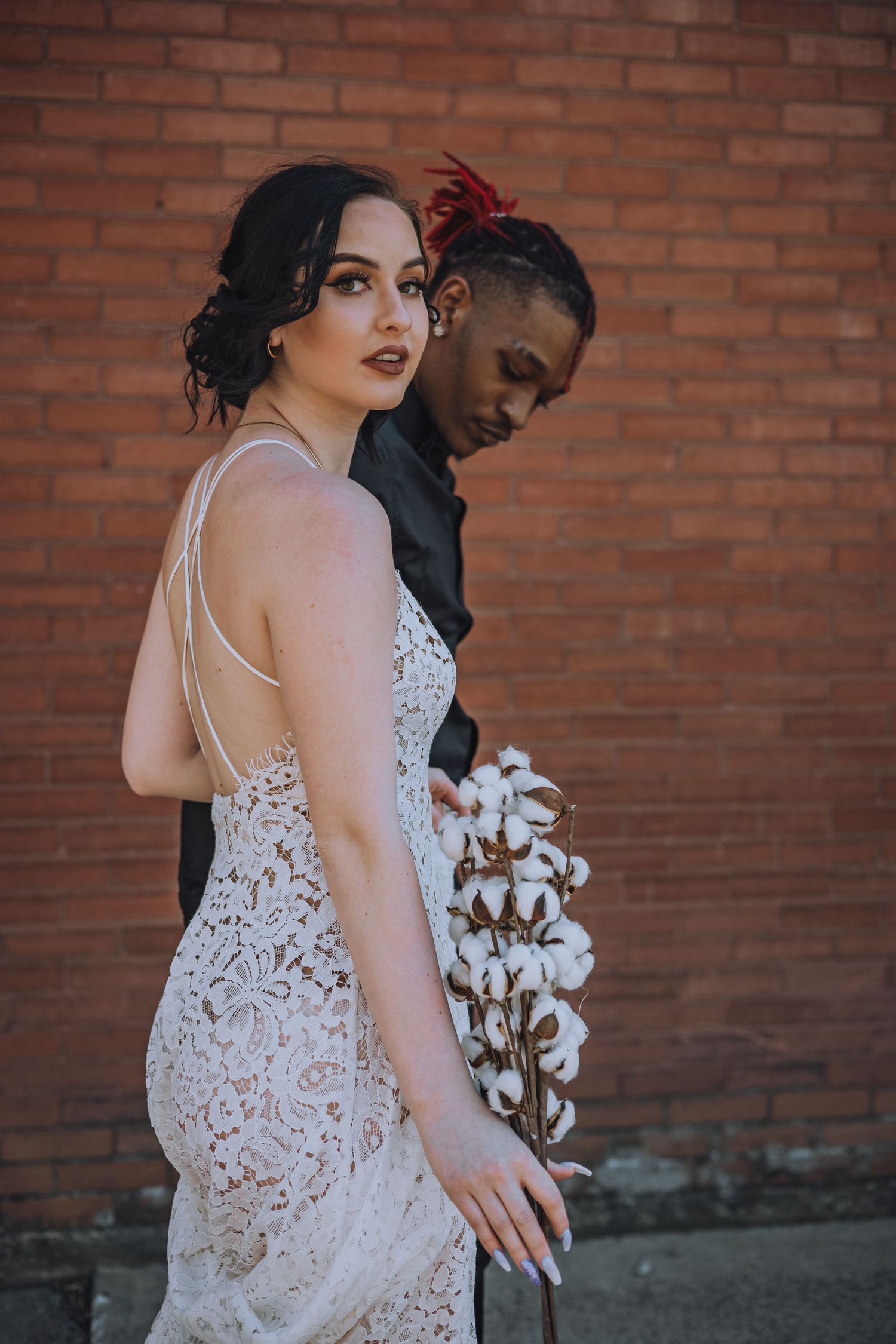 Couple in elegant attire poses against brick wall with cotton flower bouquet.