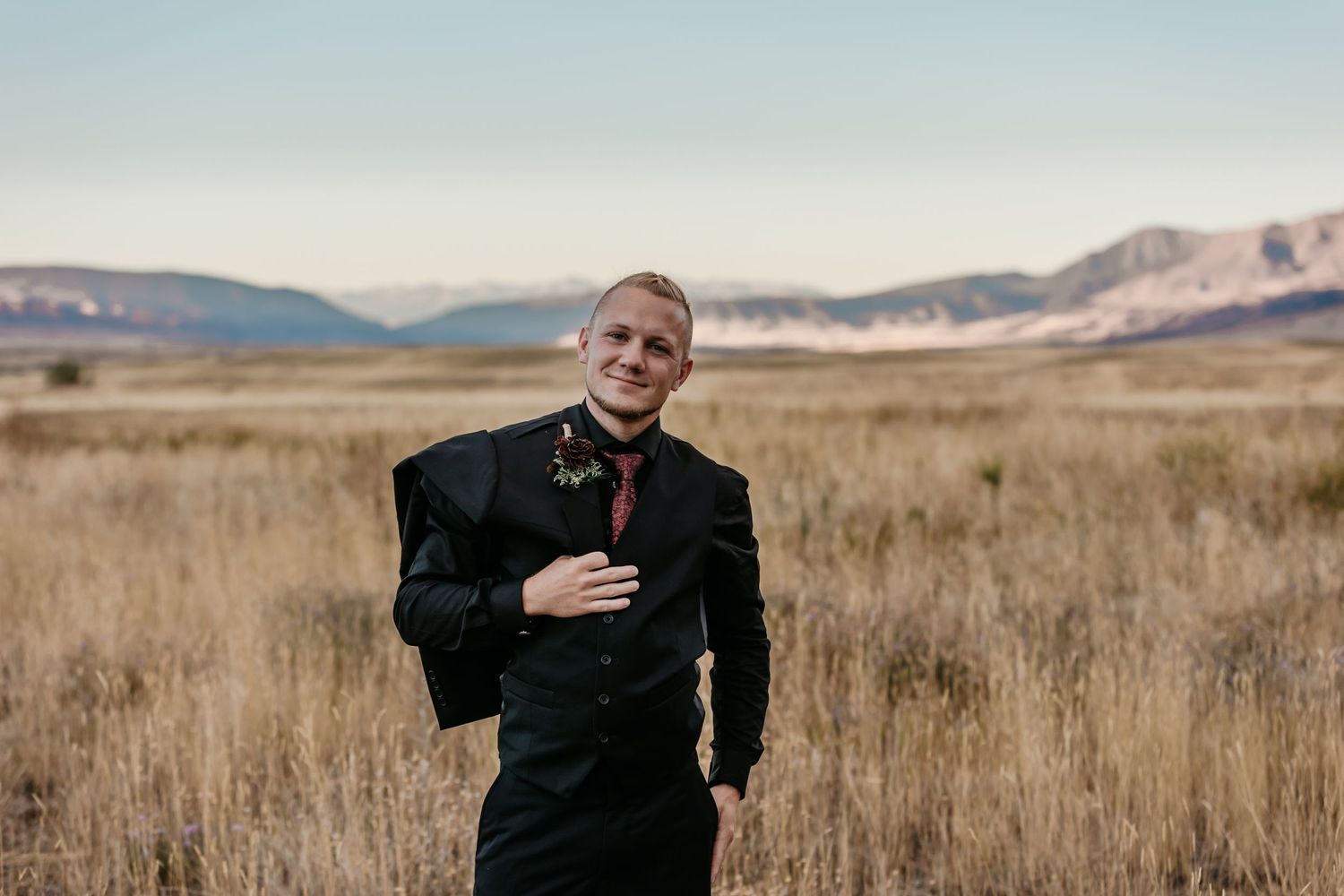 Person in black formal attire walking through golden grass field with mountains in background.