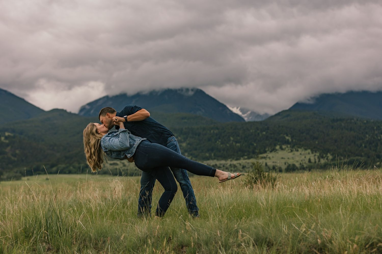 Playful couple dancing in mountain meadow with dramatic cloudy sky.
