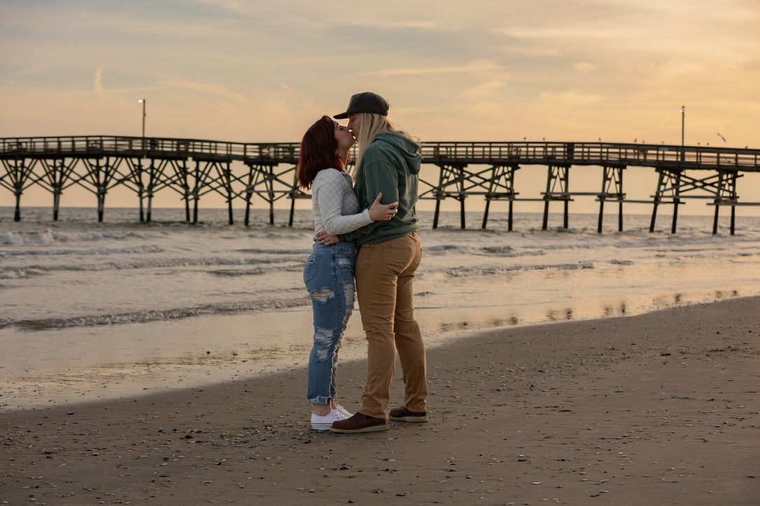 Couple embraces on beach at sunset with wooden pier in background.