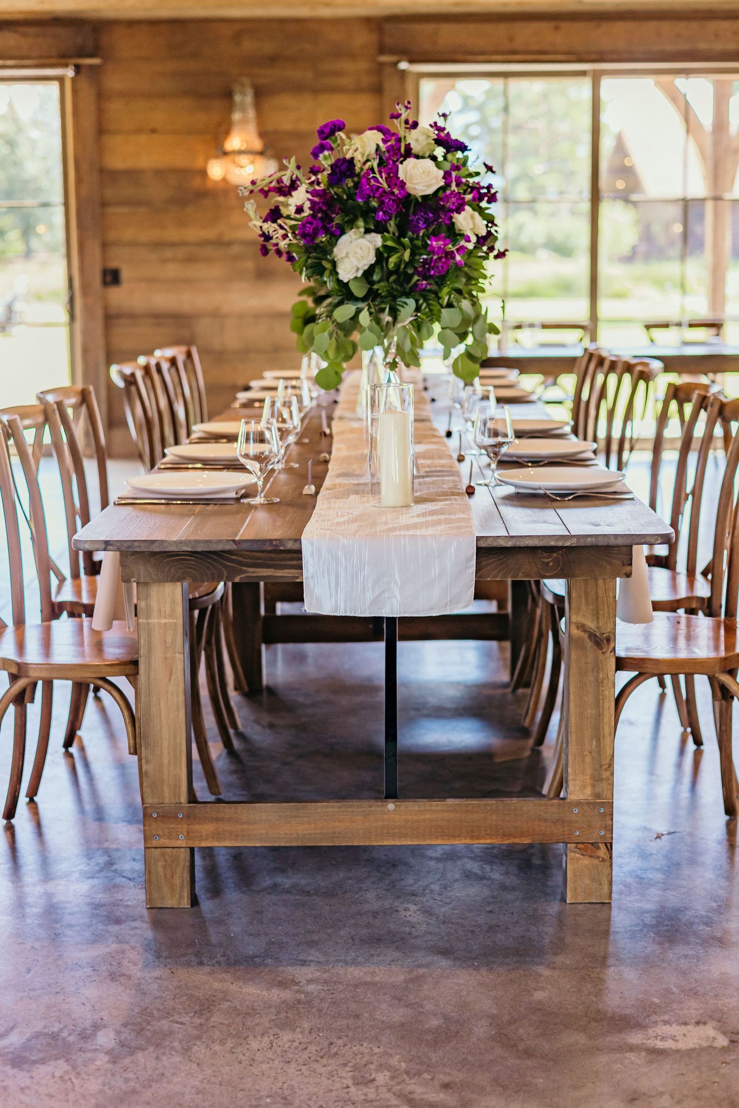 Farm tables decorated with purple floral centerpieces in a light-filled rustic reception space.
