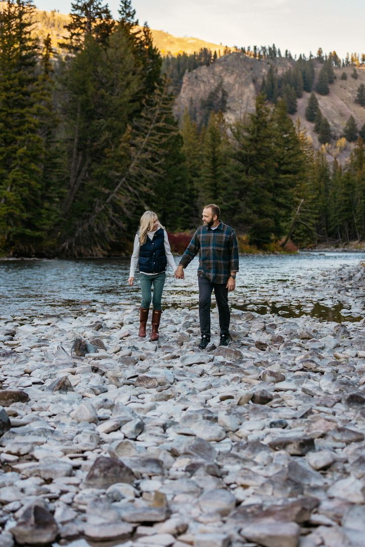 A couple walks along a rocky riverbed surrounded by evergreen trees and mountains.