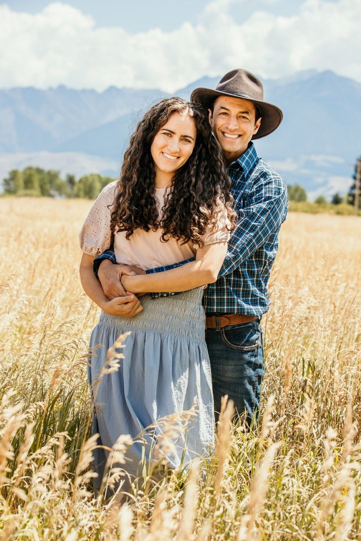 Couple shares tender moment in tall wheat field with mountain backdrop during golden hour.
