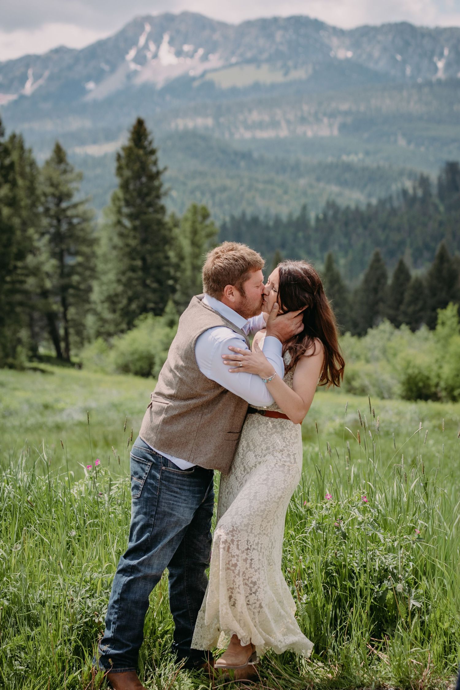 A romantic kiss in a mountain meadow with dramatic peaks and evergreen forests in the background.