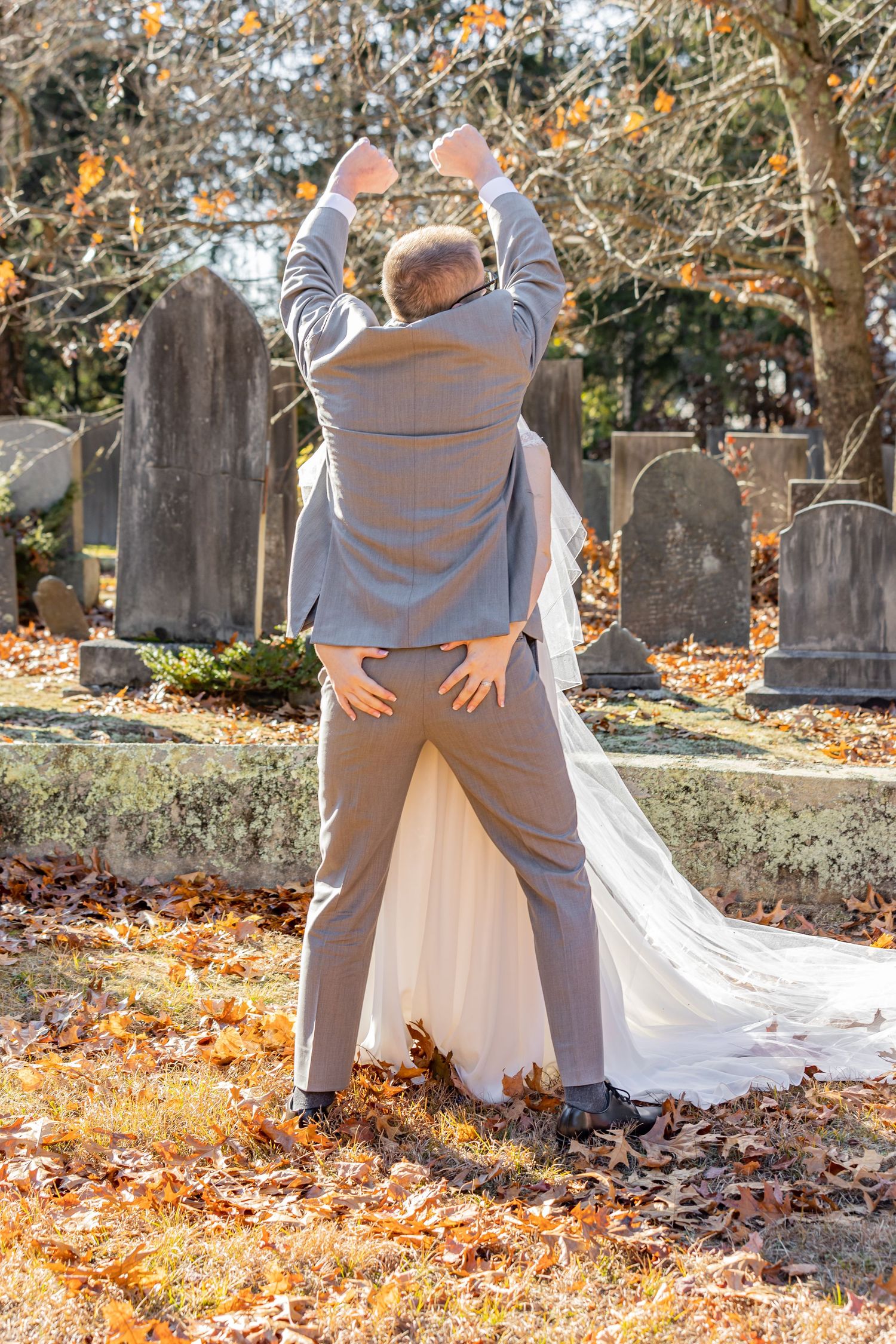 Couple playfully posing in autumn leaves at a cemetery during a wedding photoshoot.