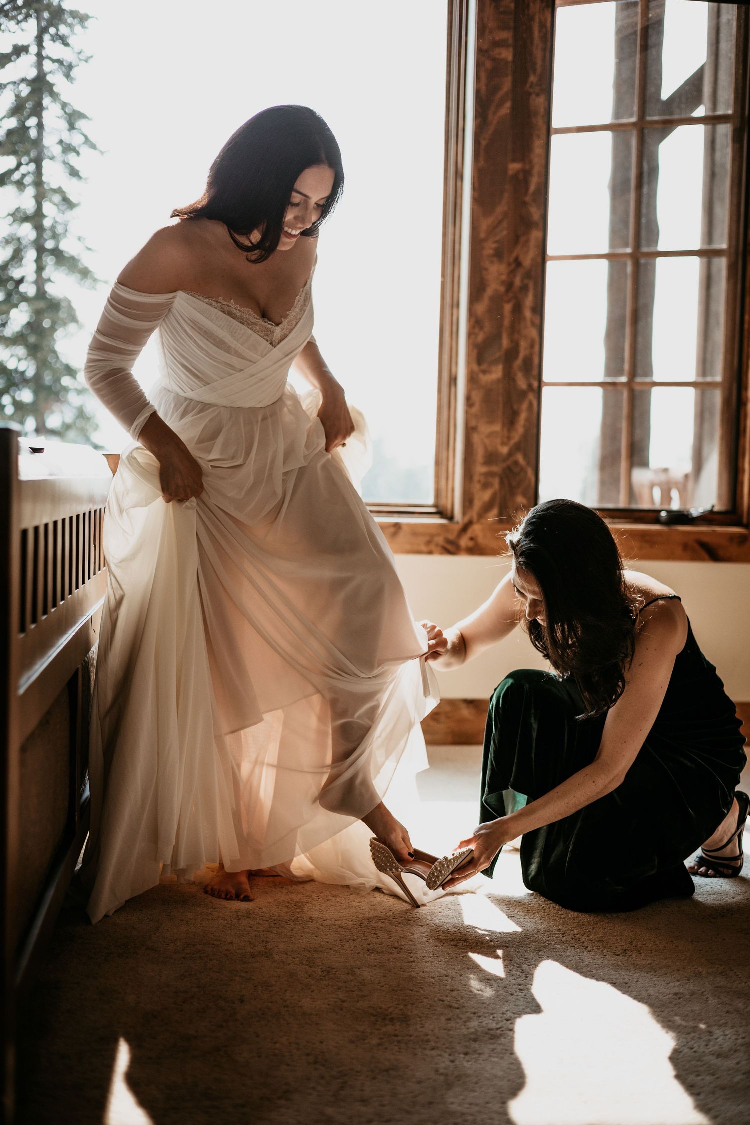 Bride in flowing white dress getting ready on rustic wooden floor with natural lighting.