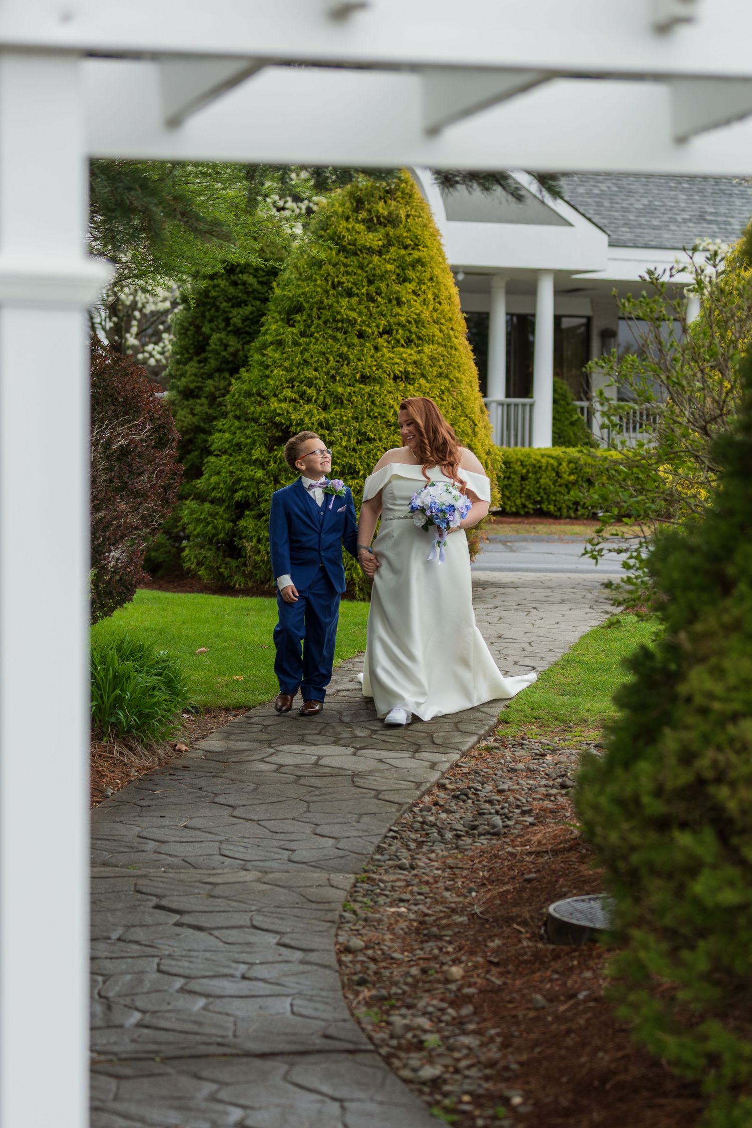 Bride and groom walk hand in hand along a stone garden path surrounded by lush greenery at an elegant wedding venue.