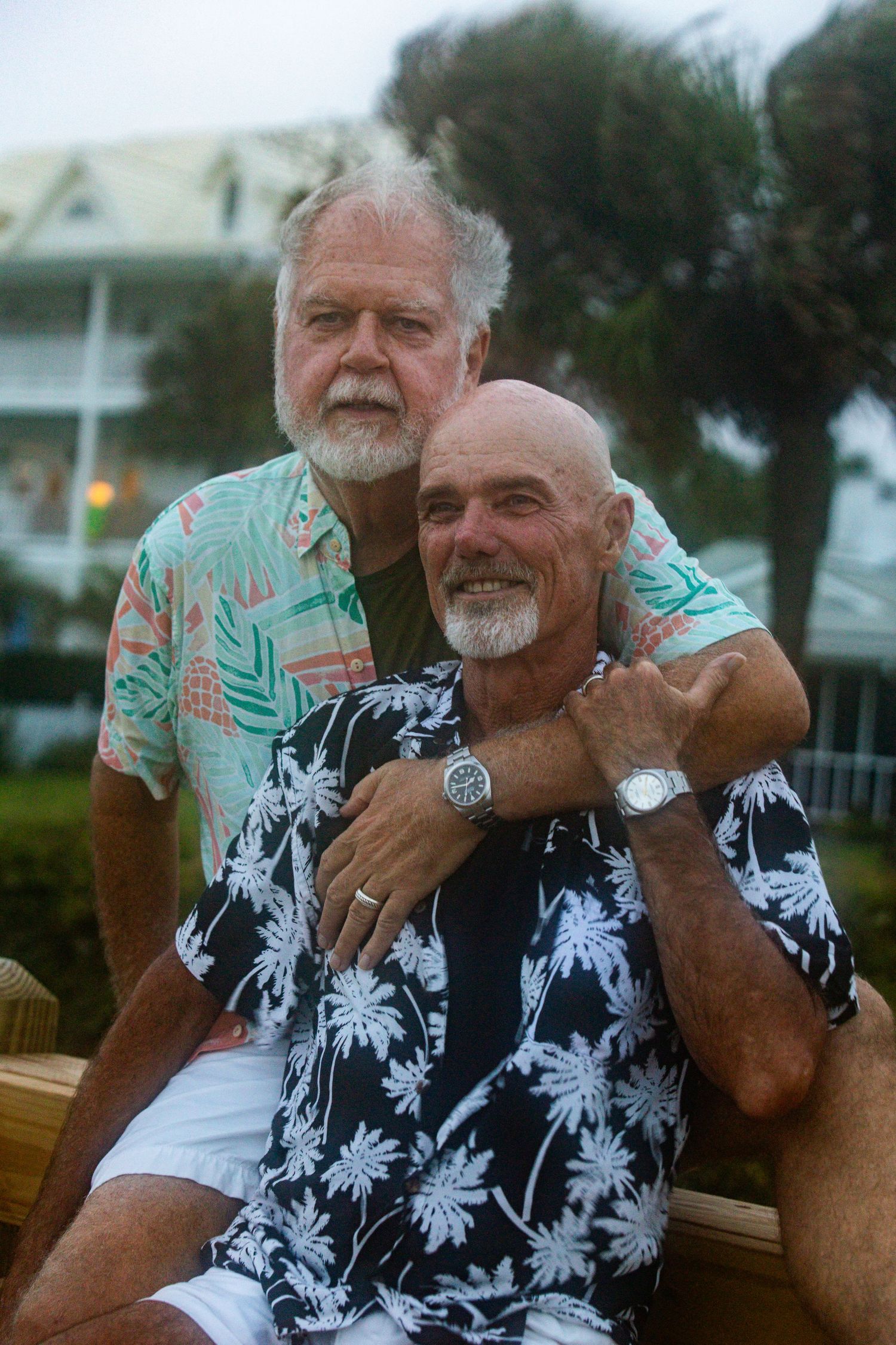 Two older men in tropical shirts pose together outdoors near palm trees, smiling warmly in a casual beach setting.