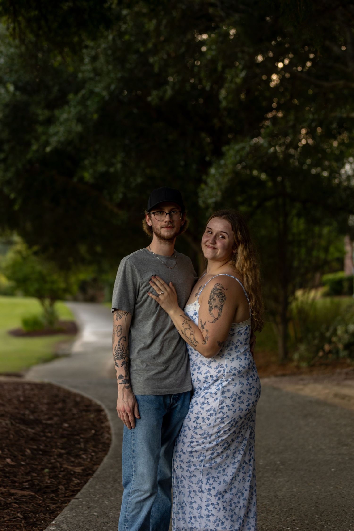 Couple with tattoos posing on a tree-lined path, woman in floral dress leaning on man in gray tee.