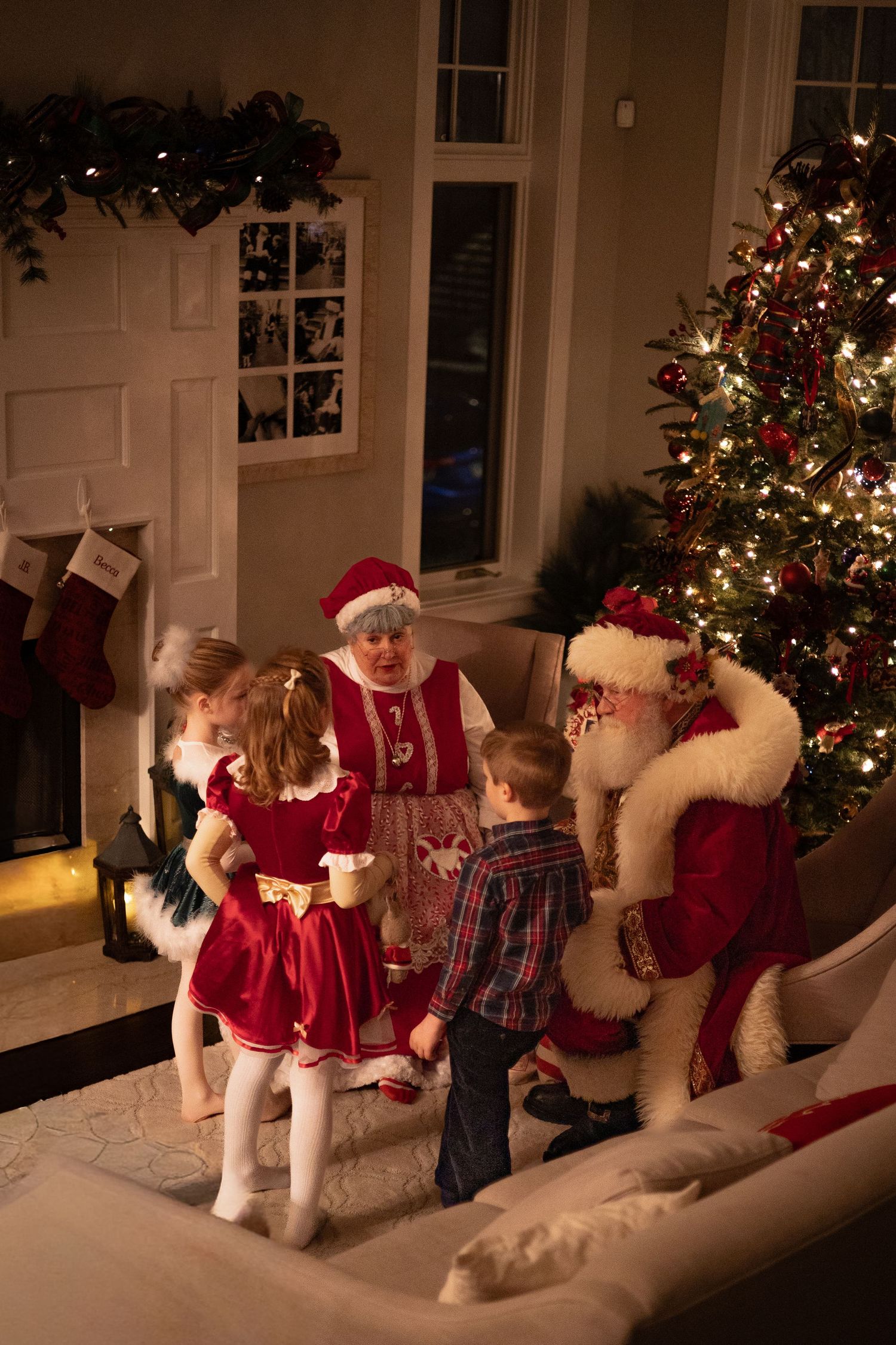 Children meet Santa and Mrs. Claus by a decorated Christmas tree and fireplace in a cozy holiday living room.