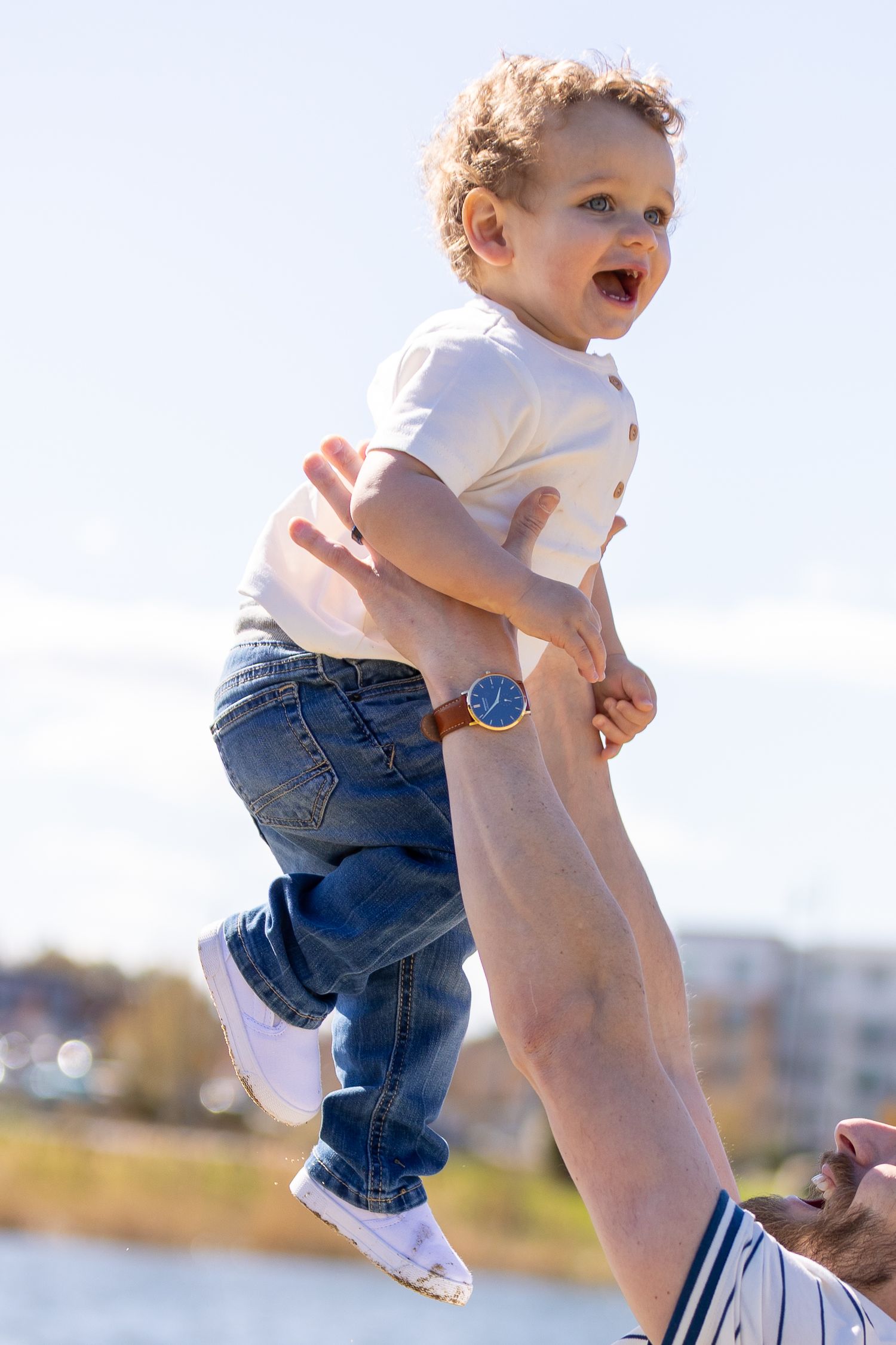 A joyful toddler in jeans and white shirt is lifted high in the air by an adult outdoors on a sunny day.