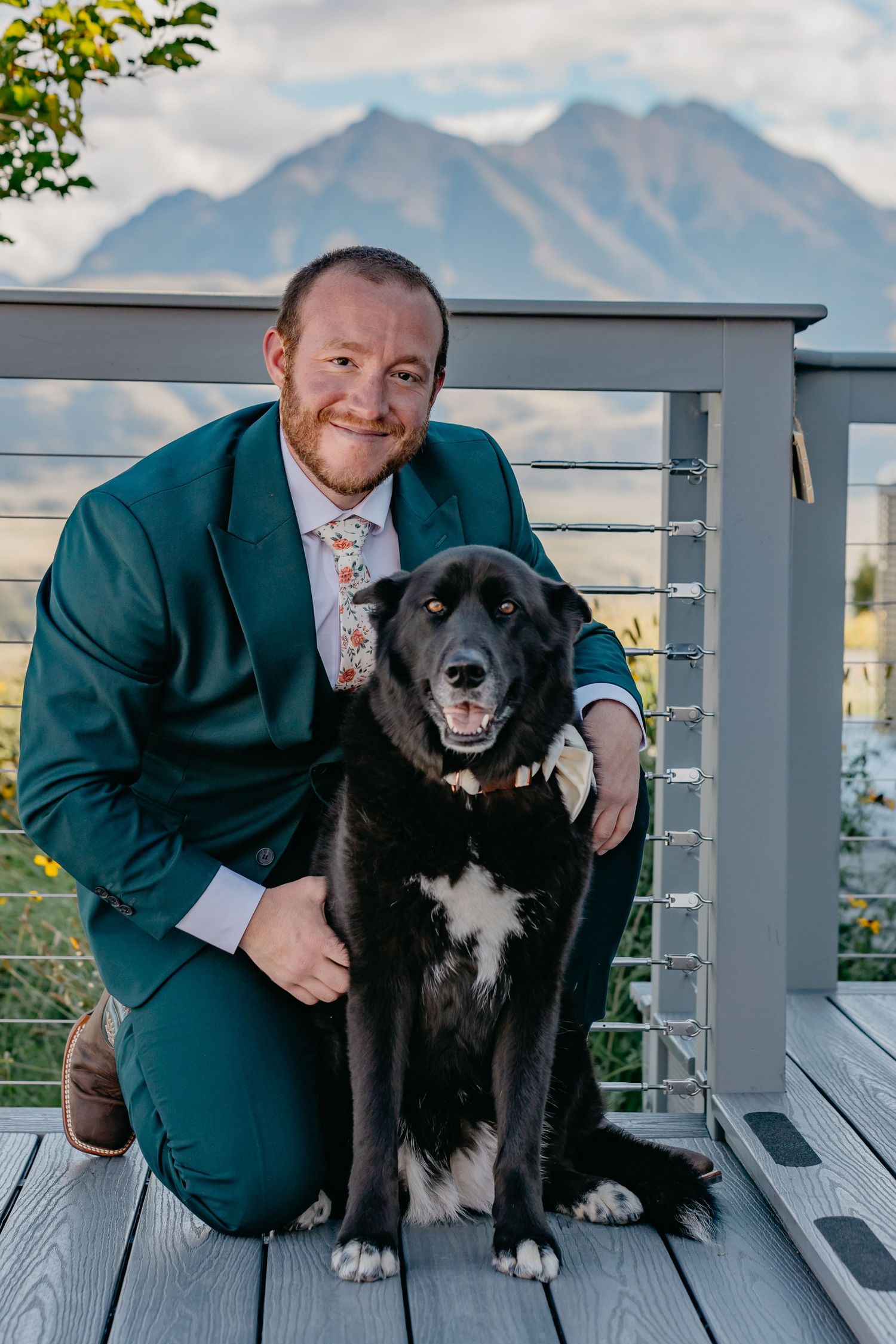 Black and white dog posing with person in teal suit on deck with mountain views.