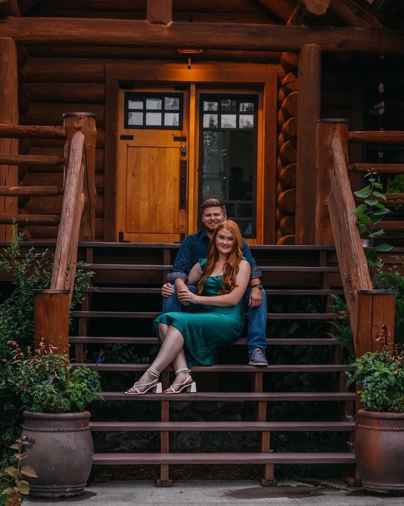 A couple relaxes on wooden cabin steps surrounded by potted plants and rustic architecture.