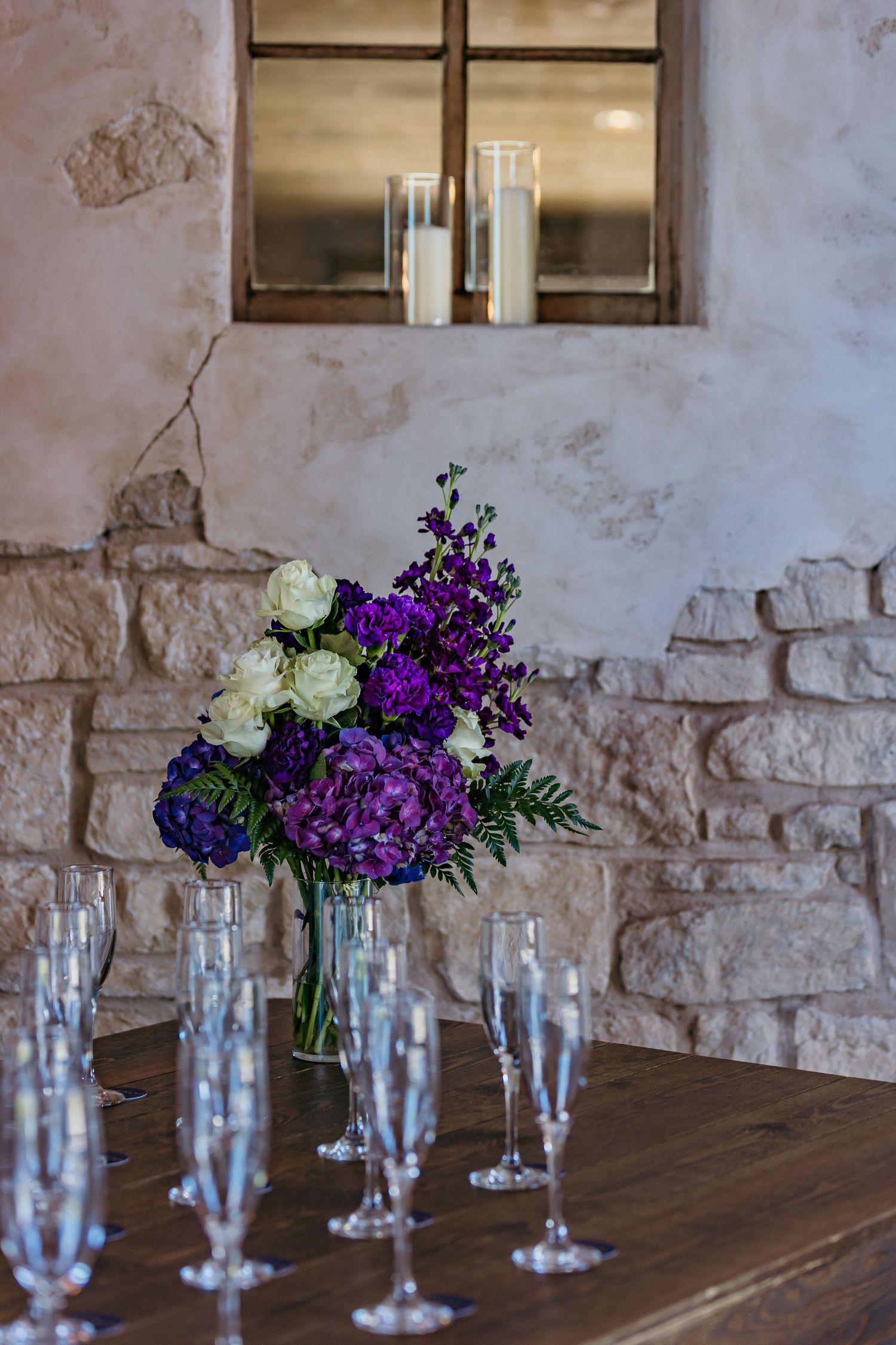 Purple and white floral arrangements in glass vases line a rustic wooden table.