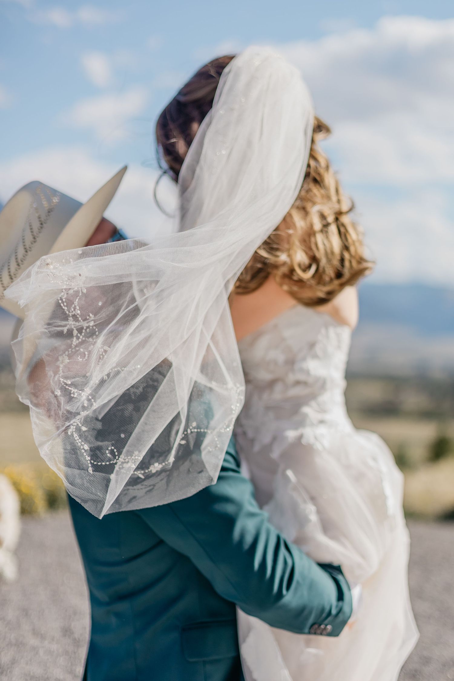 A romantic wedding moment captured against a scenic mountain backdrop with flowing veil and teal jacket details.