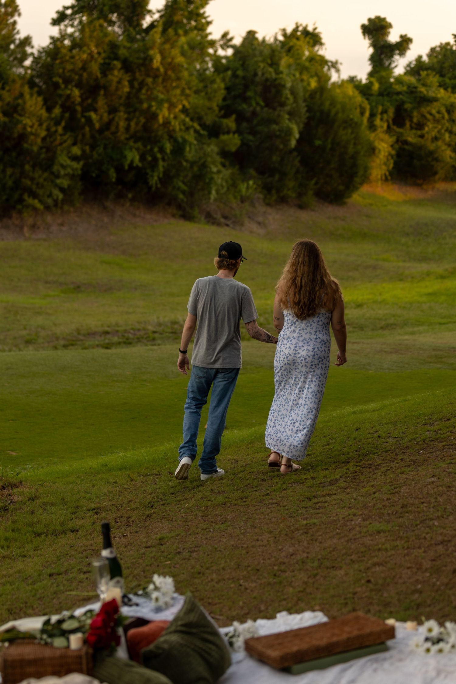 Couple holding hands walking on green grass at sunset with picnic setup in foreground.