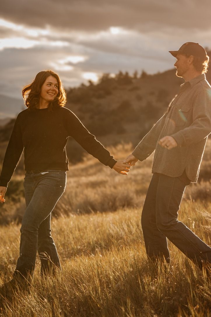 Couple running playfully through tall grass field at golden hour.