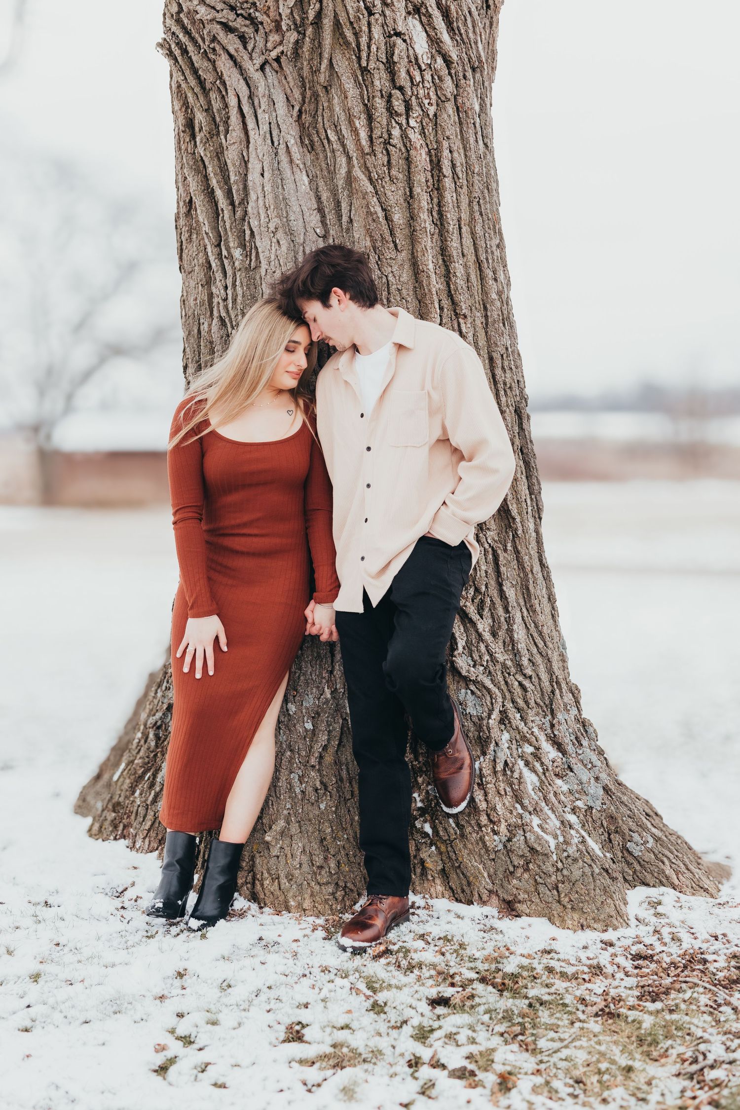 A romantic winter engagement photo with a couple sitting by a large tree trunk in the snow.