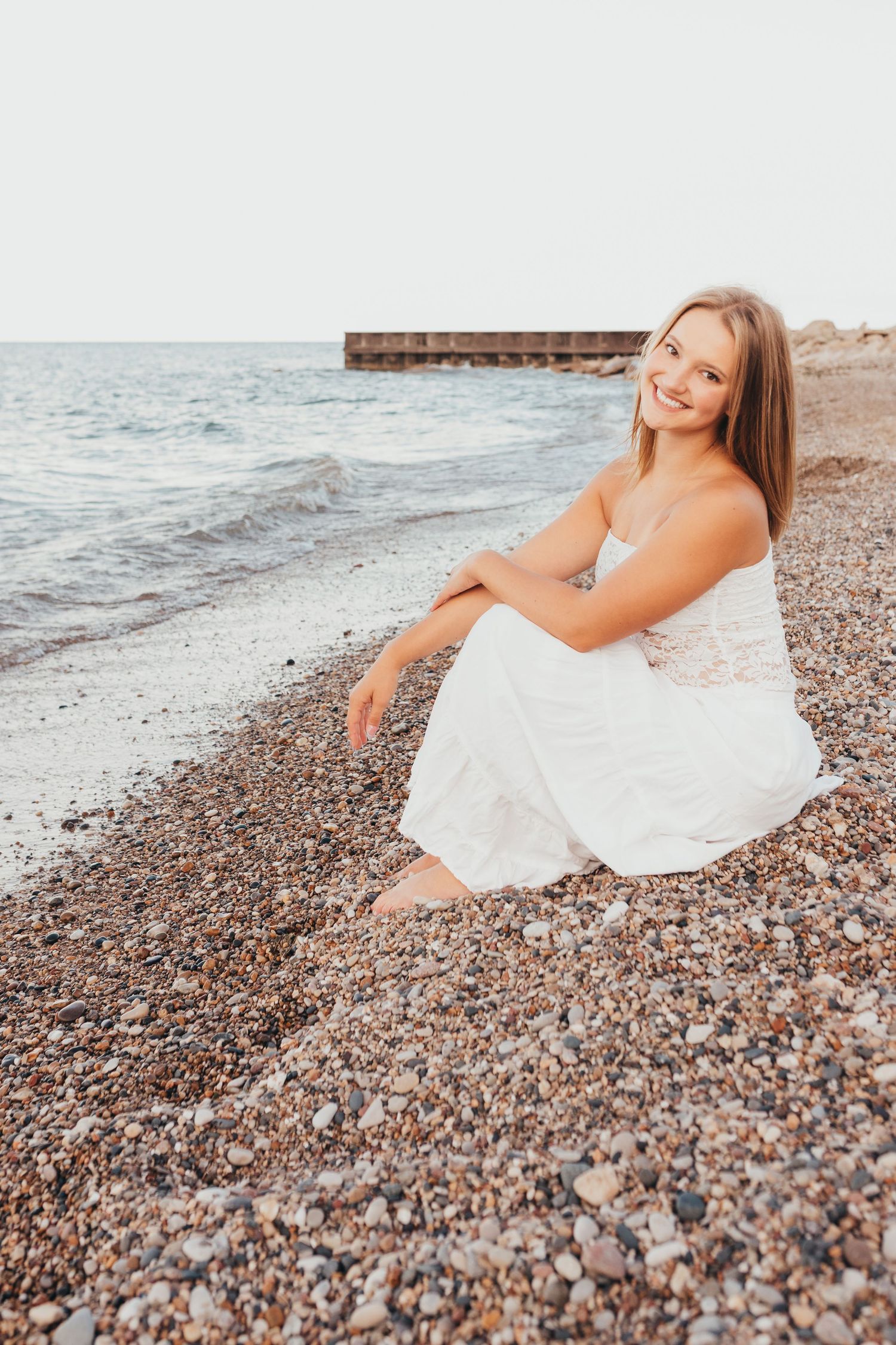 A dreamy beach photoshoot features a flowing white dress on a pebbled shoreline.