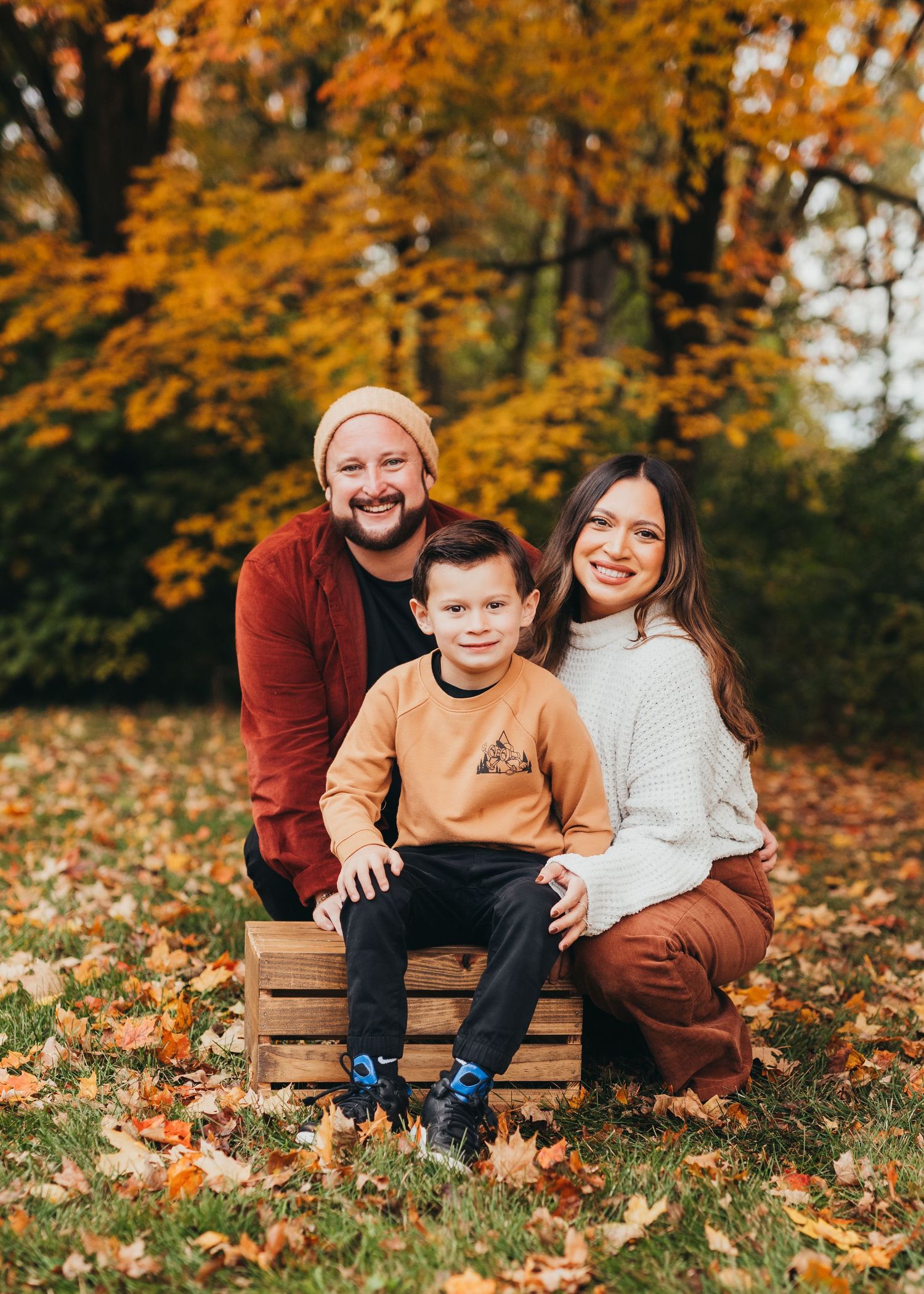 Family sits together on wooden bench surrounded by vibrant autumn leaves in an outdoor setting.