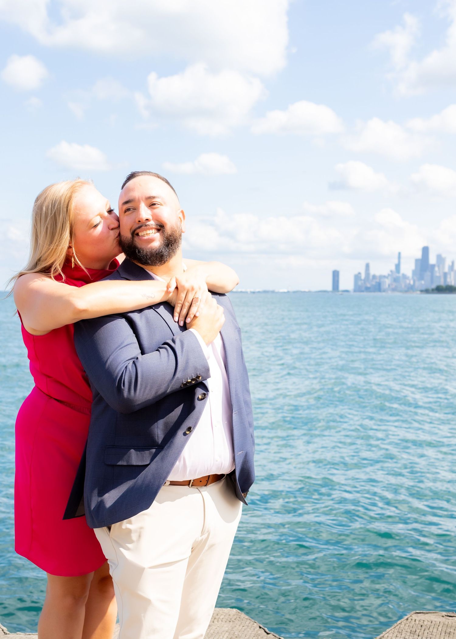 A couple embraces joyfully by the waterfront with a city skyline visible in the background.