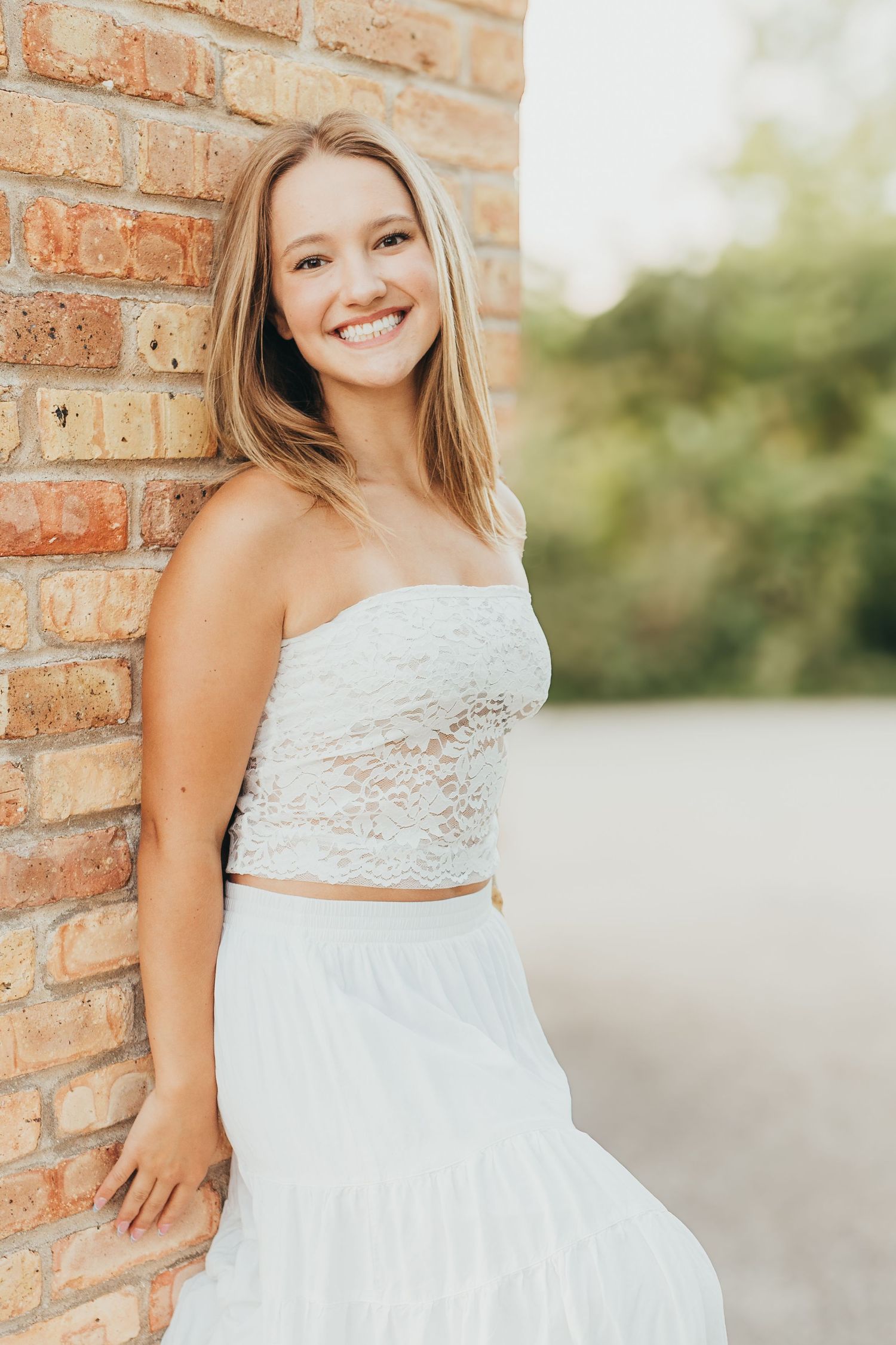 A joyful portrait against a brick wall wearing a white lace top and flowing skirt.