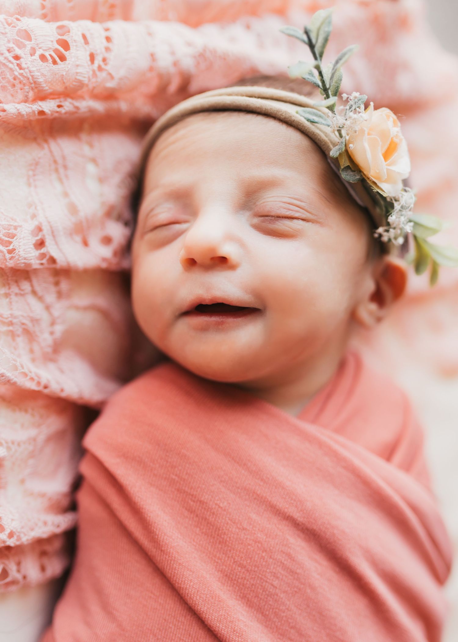 Newborn baby wrapped in pink blanket wearing floral headband smiling peacefully.
