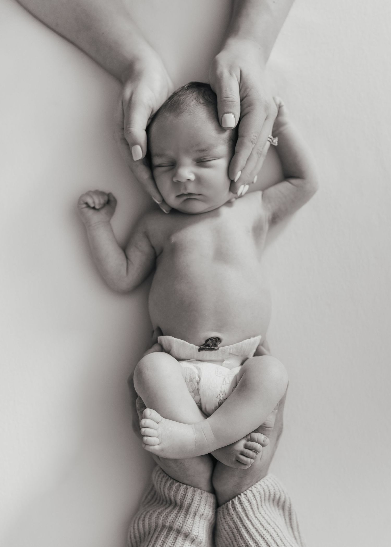 A peaceful black and white portrait of a newborn baby lying on a soft surface with gentle hands cradling their head.