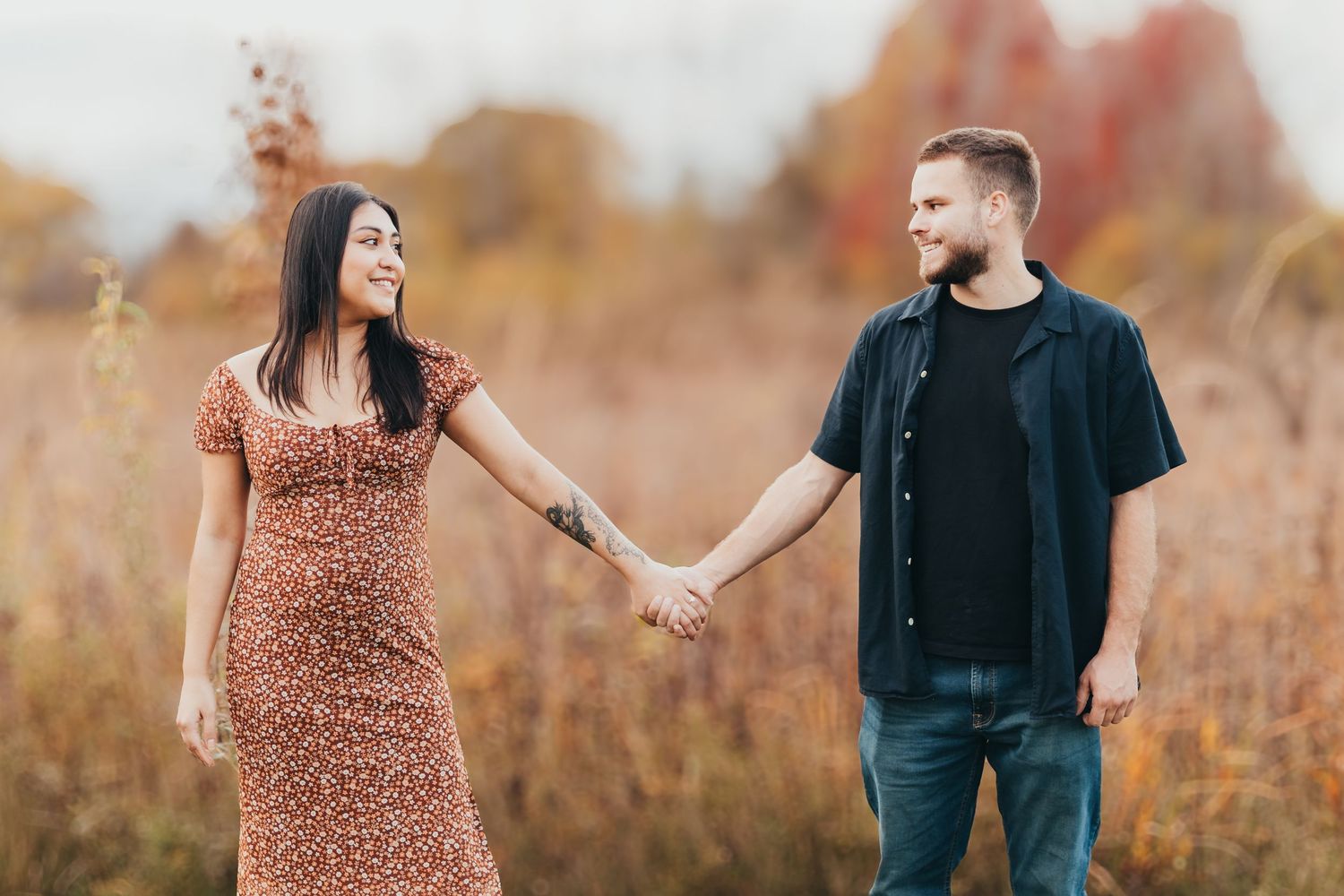 A couple in autumn attire walks hand in hand through a field of tall grass.