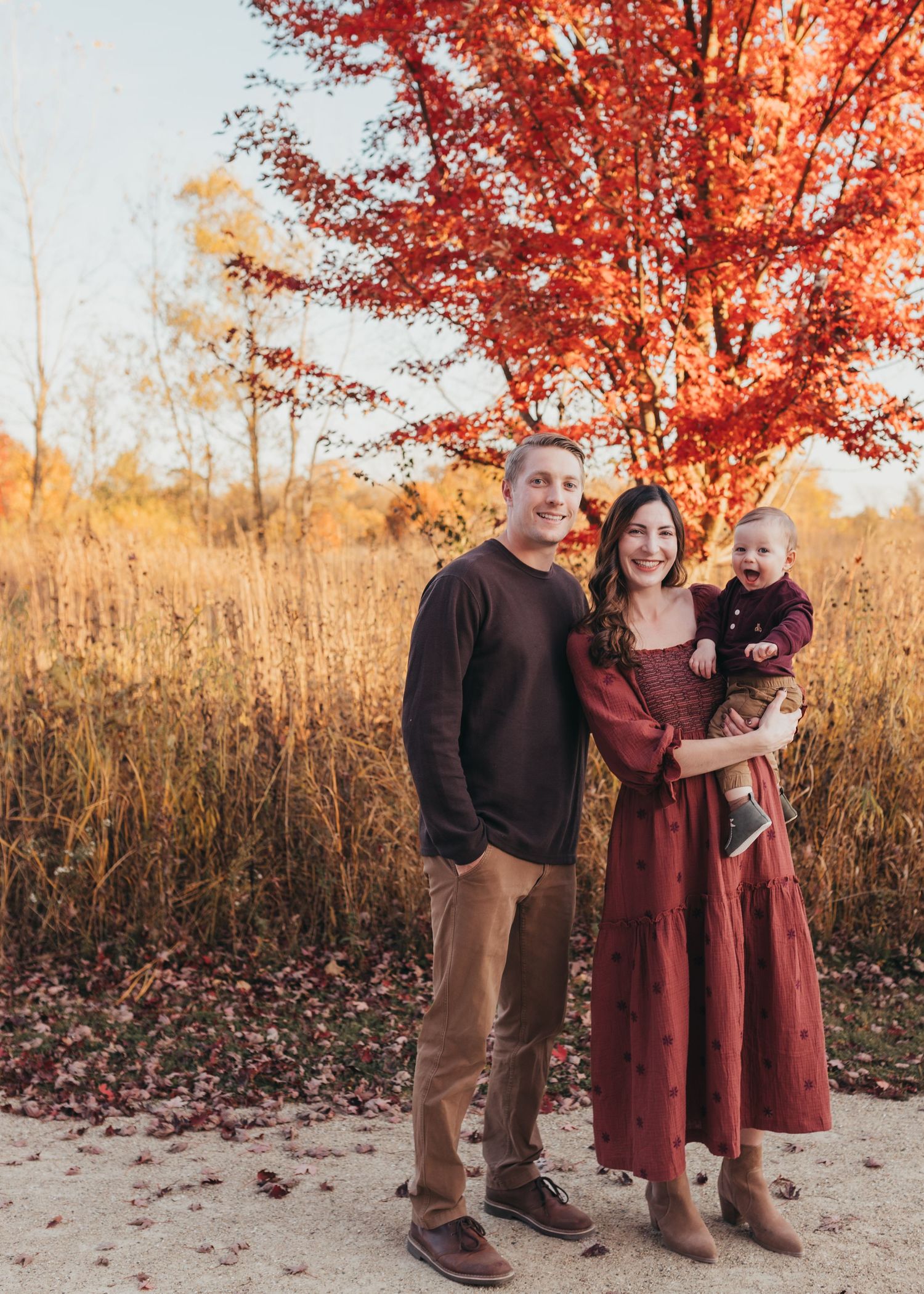 A family stands together against vibrant fall foliage with red maple leaves and golden grass in the background.