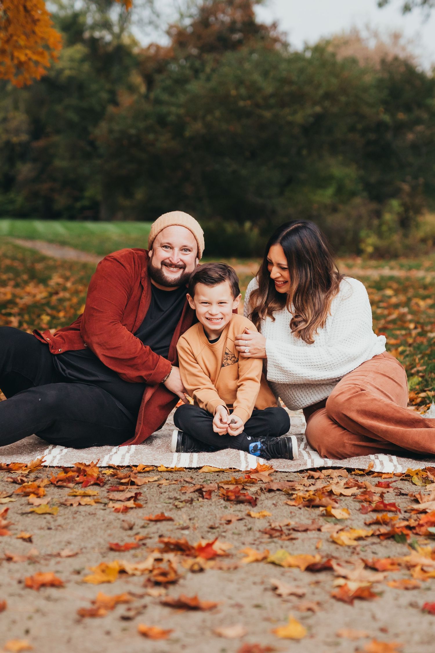 Family lying together on blanket surrounded by fallen autumn leaves in outdoor park setting.