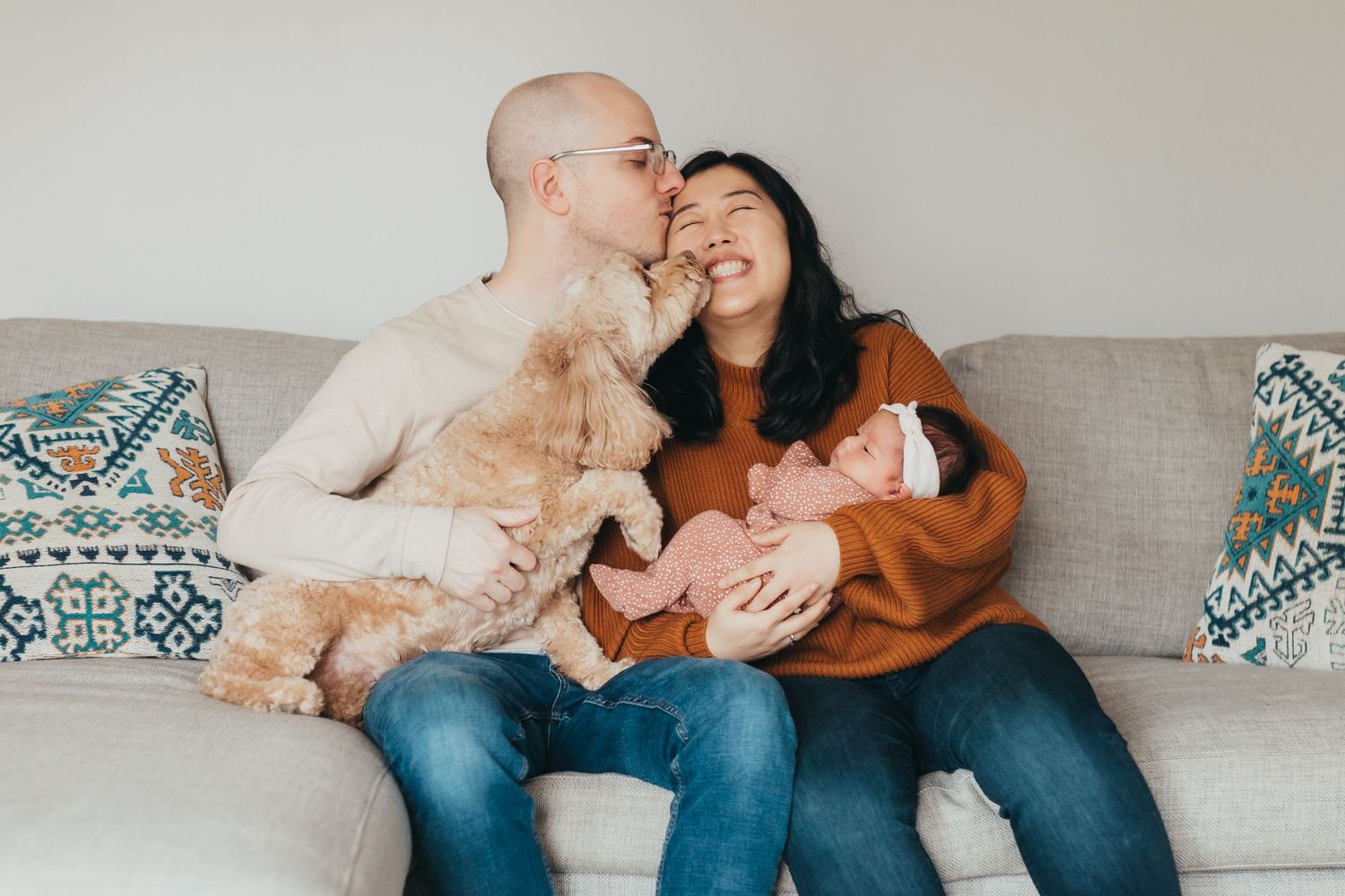 Couple cuddling on couch with their dog while holding a baby.
