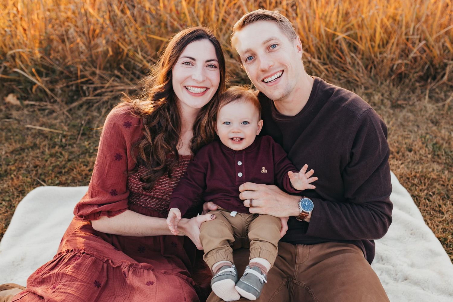 A family sits together on a white blanket surrounded by golden autumn grass during sunset.
