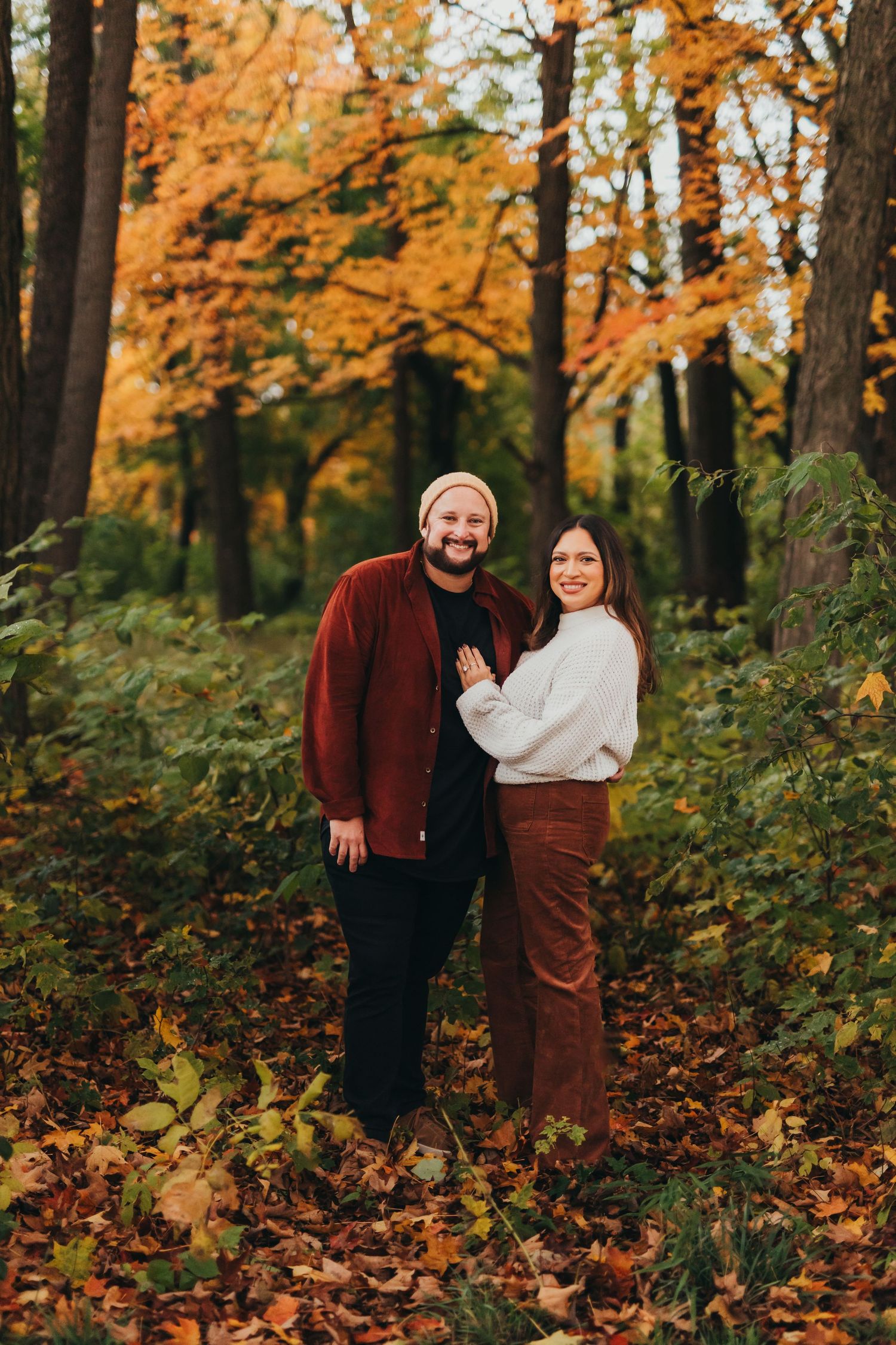 A couple stands together in a forest filled with vibrant orange autumn foliage and fallen leaves.