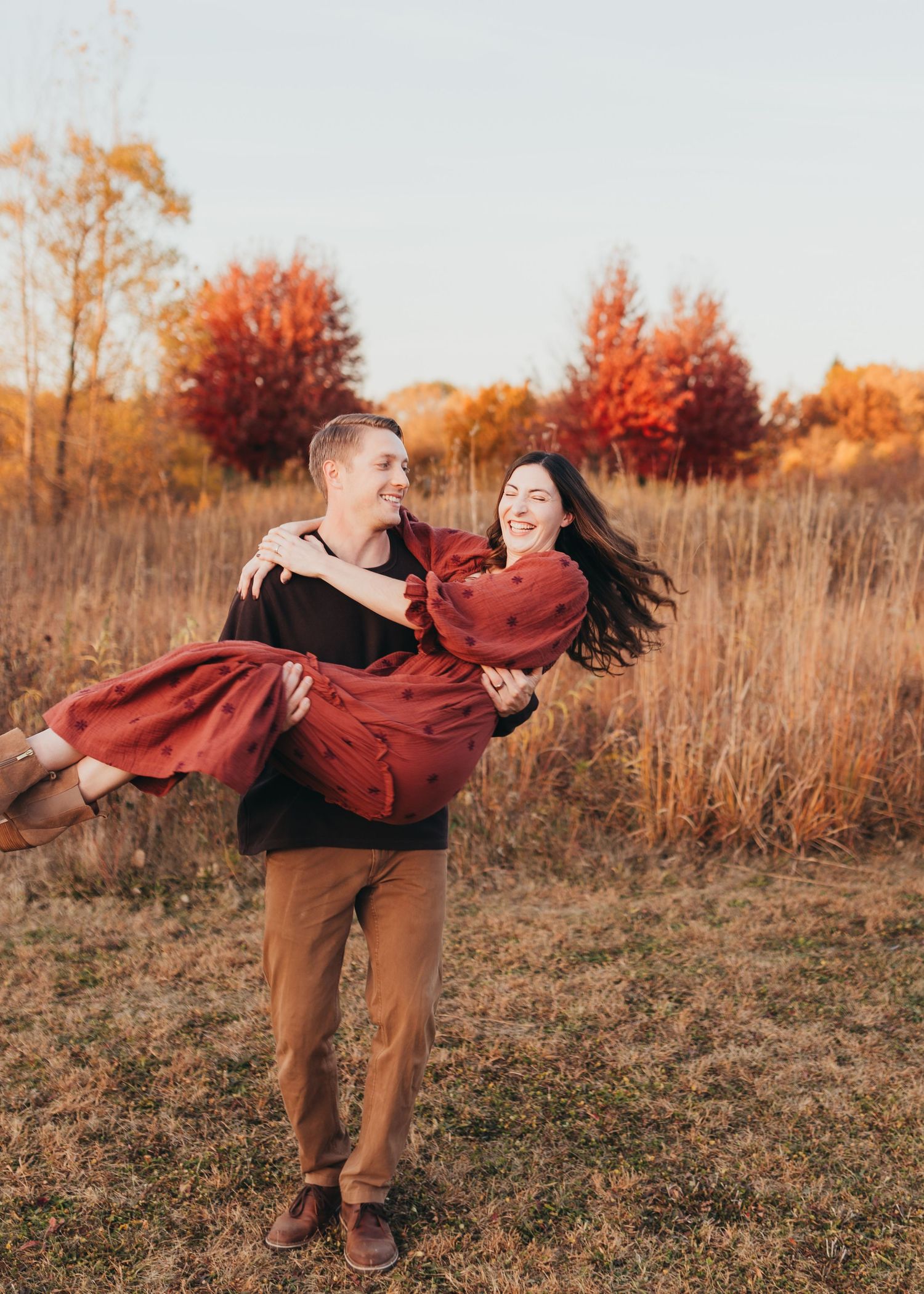 Two people dance and twirl playfully in a golden autumn field at sunset with red maple trees in the background.