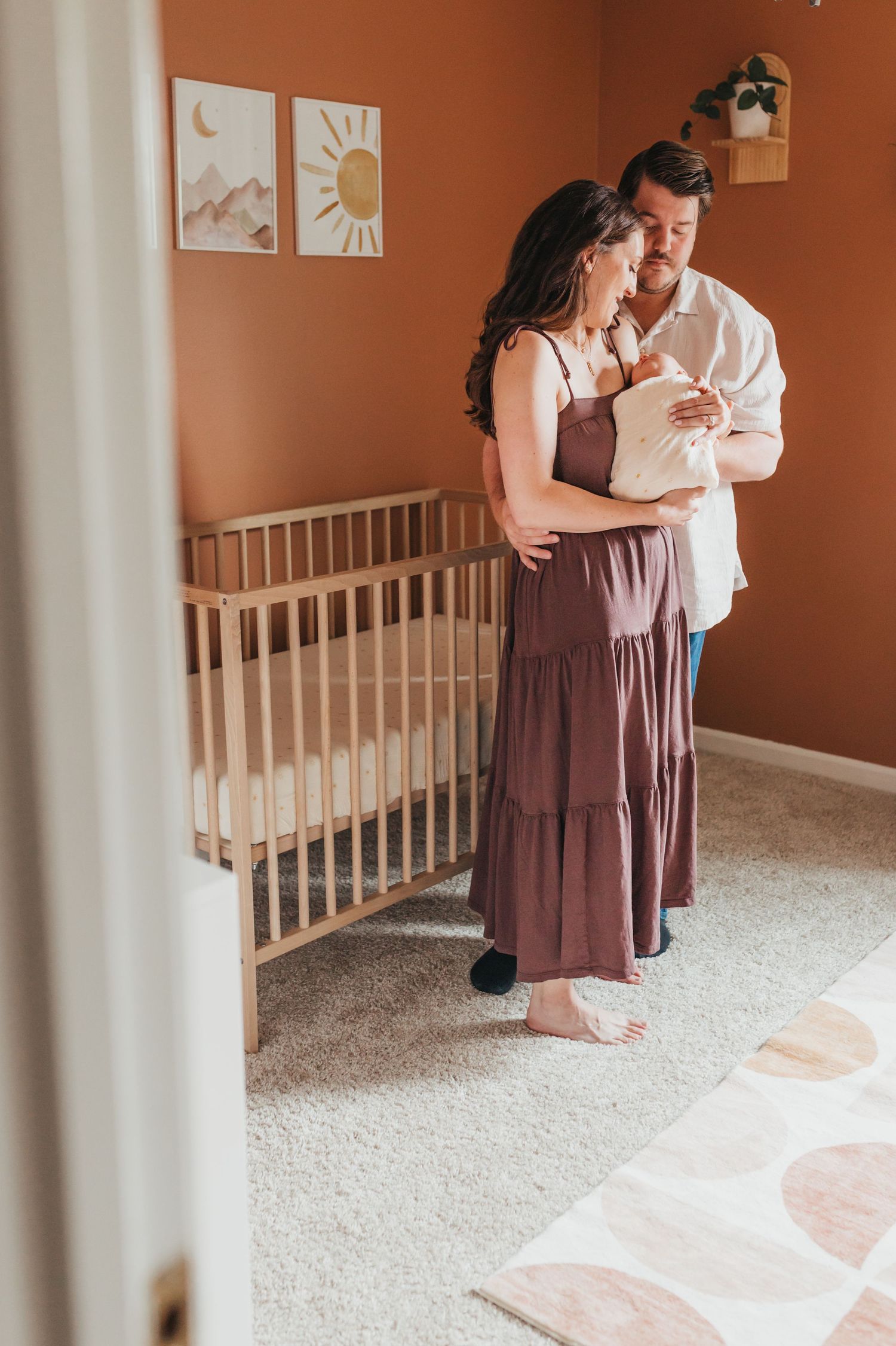 A couple embraces lovingly in a nursery room with warm terracotta walls and a wooden crib.
