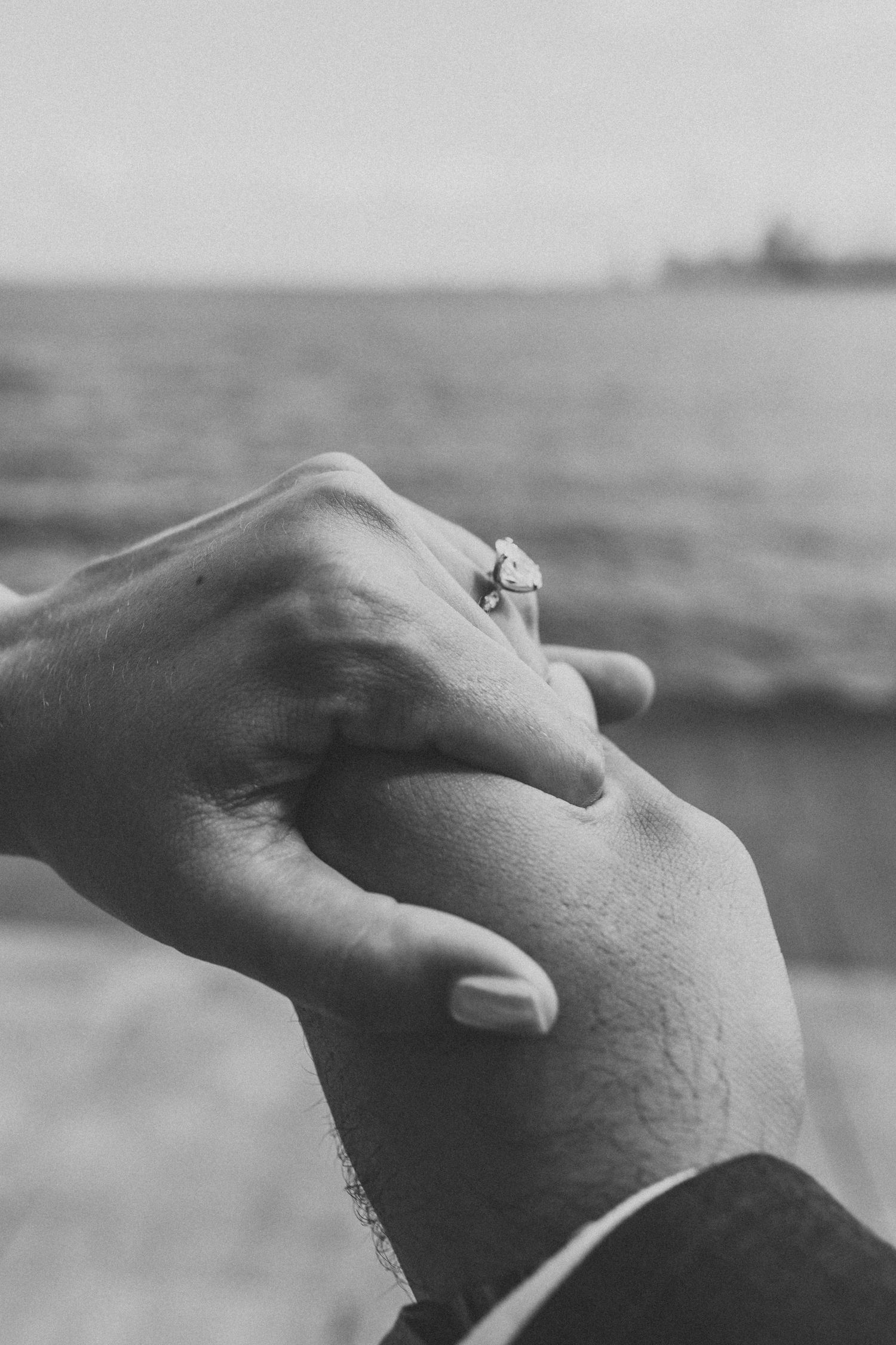 Close-up black and white photo of an engagement ring on a hand with ocean in background.