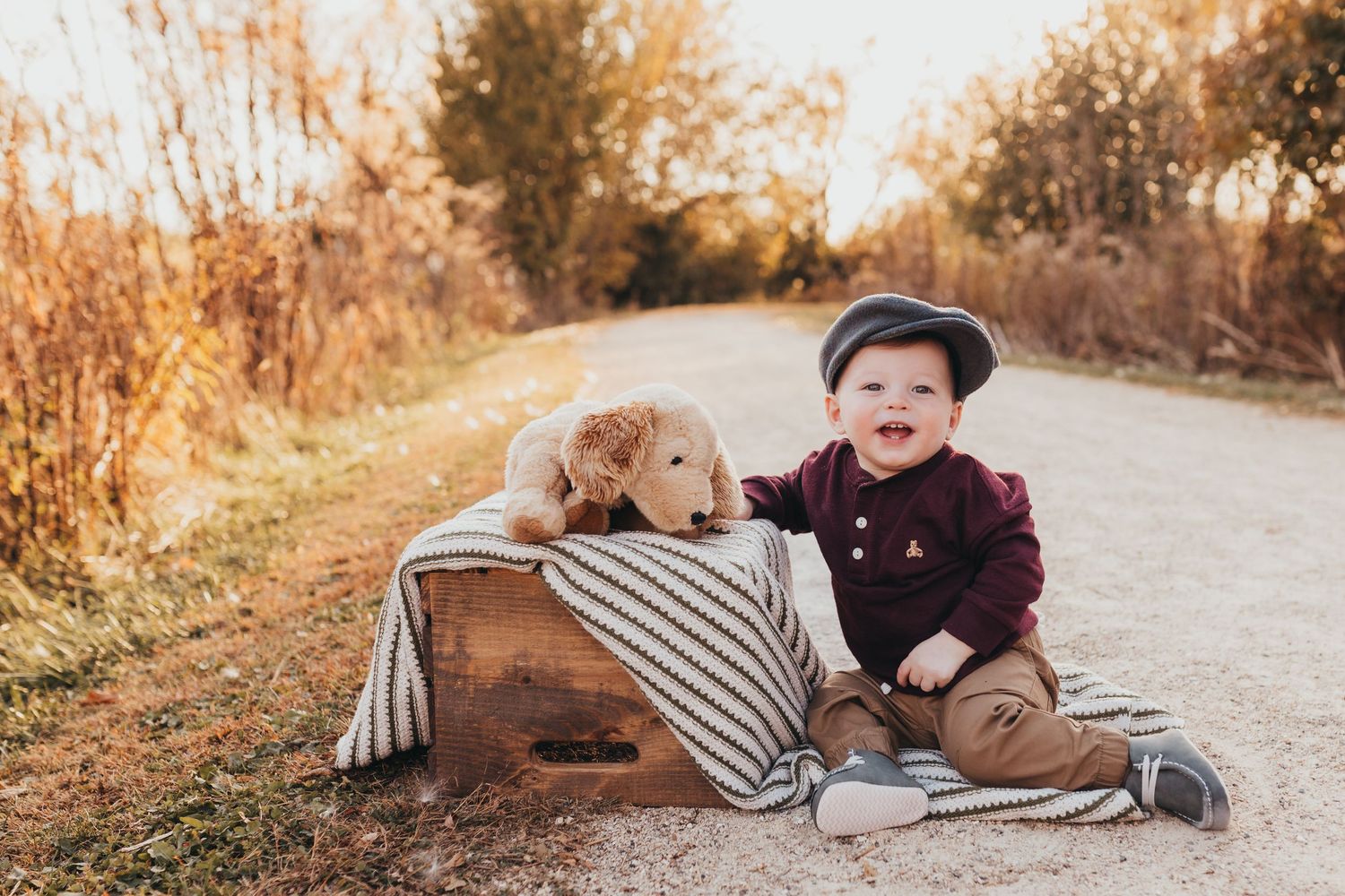 A baby in a brown sweater lies on a striped blanket during a fall photoshoot on a dirt path.