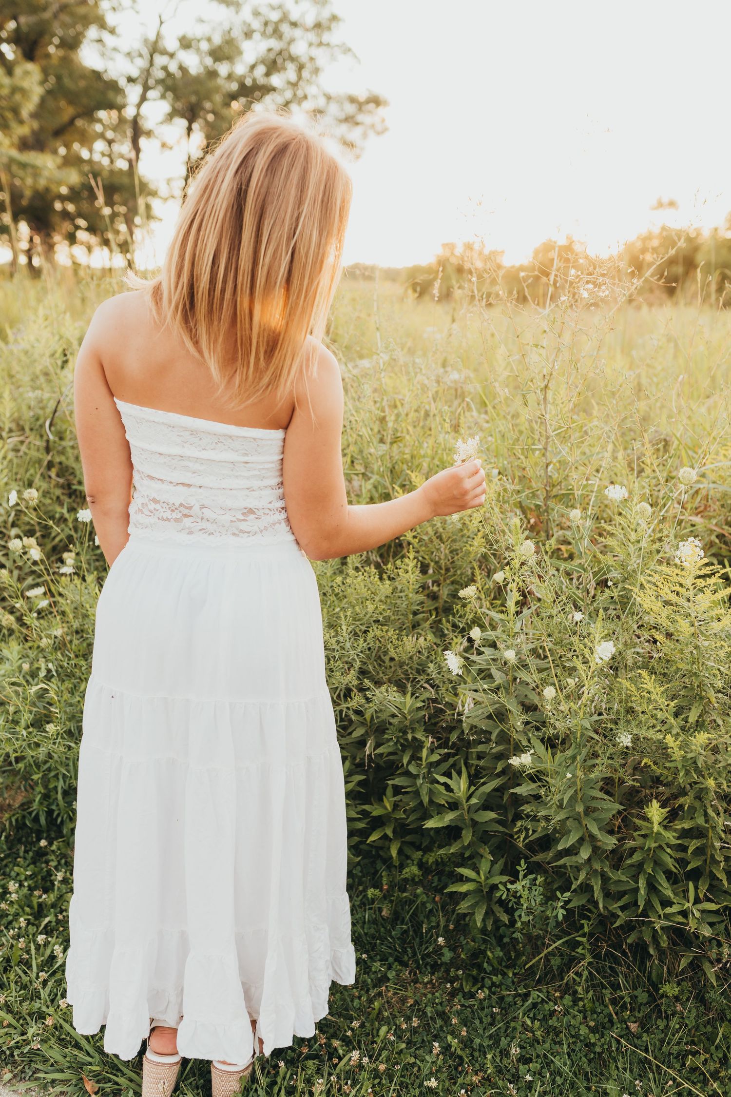 A back view of a woman in a white dress standing among wildflowers at sunset.