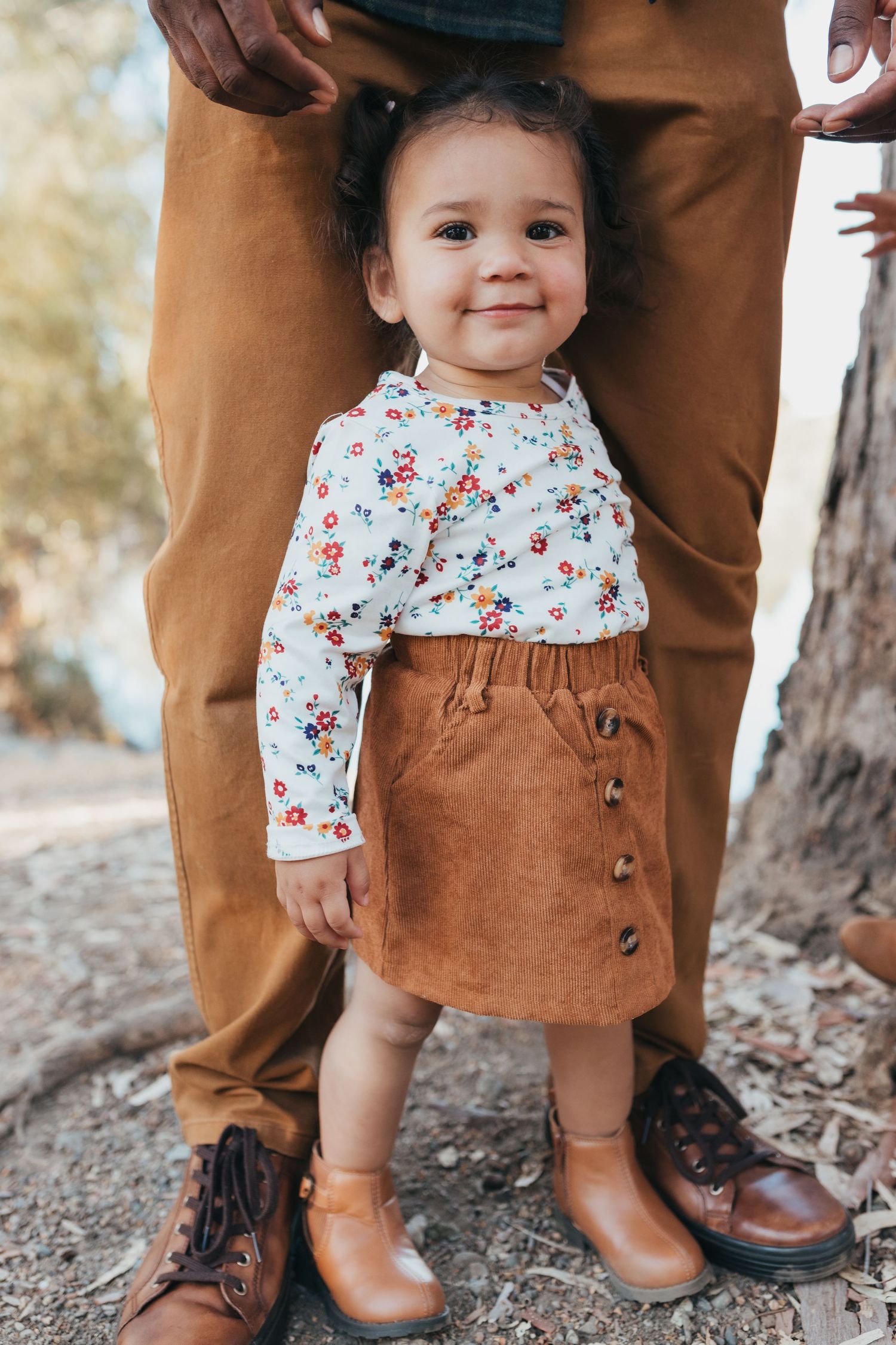 Small child wearing a floral top and brown corduroy skirt stands between adult legs in matching brown pants and boots.